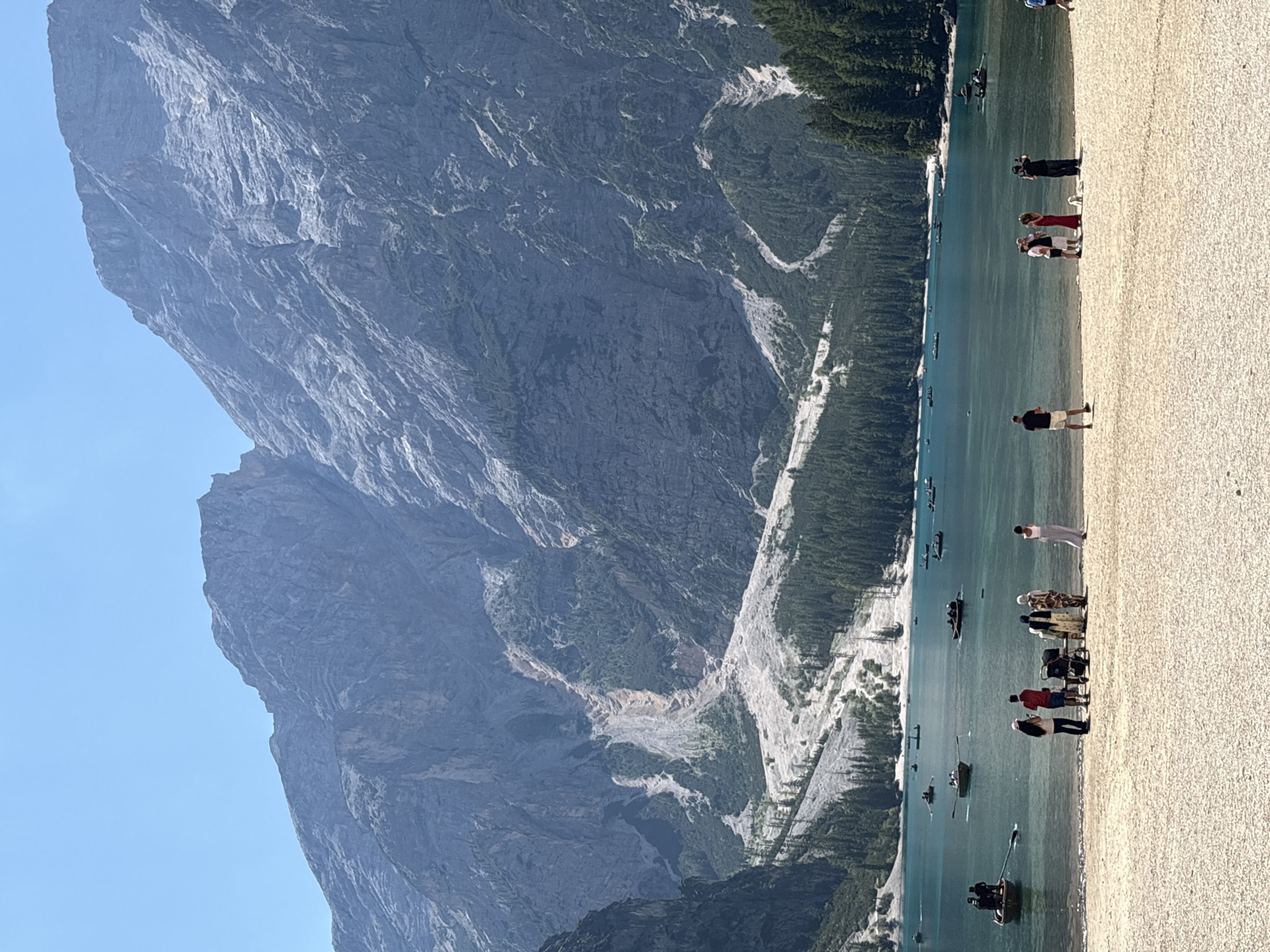 The image depicts a scenic view of Pragser Wildsee (Lago di Braies) in Prags, Italy, on a clear day. The lake is a vibrant turquoise color and dotted with numerous small boats, many of which are being rowed by people. The shoreline is a light-colored gravel beach.
In the foreground, several groups of people are visible on the beach. One group, on the left, consists of four individuals, two of whom appear to be looking out at the lake. Next to them, a family stands near a stroller. Further to the right, another group includes a woman in a long, flowing dress, and a man dressed in black holding a camera, possibly taking photos.
The background is dominated by the imposing mountains that surround the lake. The mountains are rocky and steep, with some areas showing signs of erosion and scree slopes. Patches of green vegetation, including pine trees, cling to the lower slopes and the edges of the lake. The sky is a clear, bright blue.