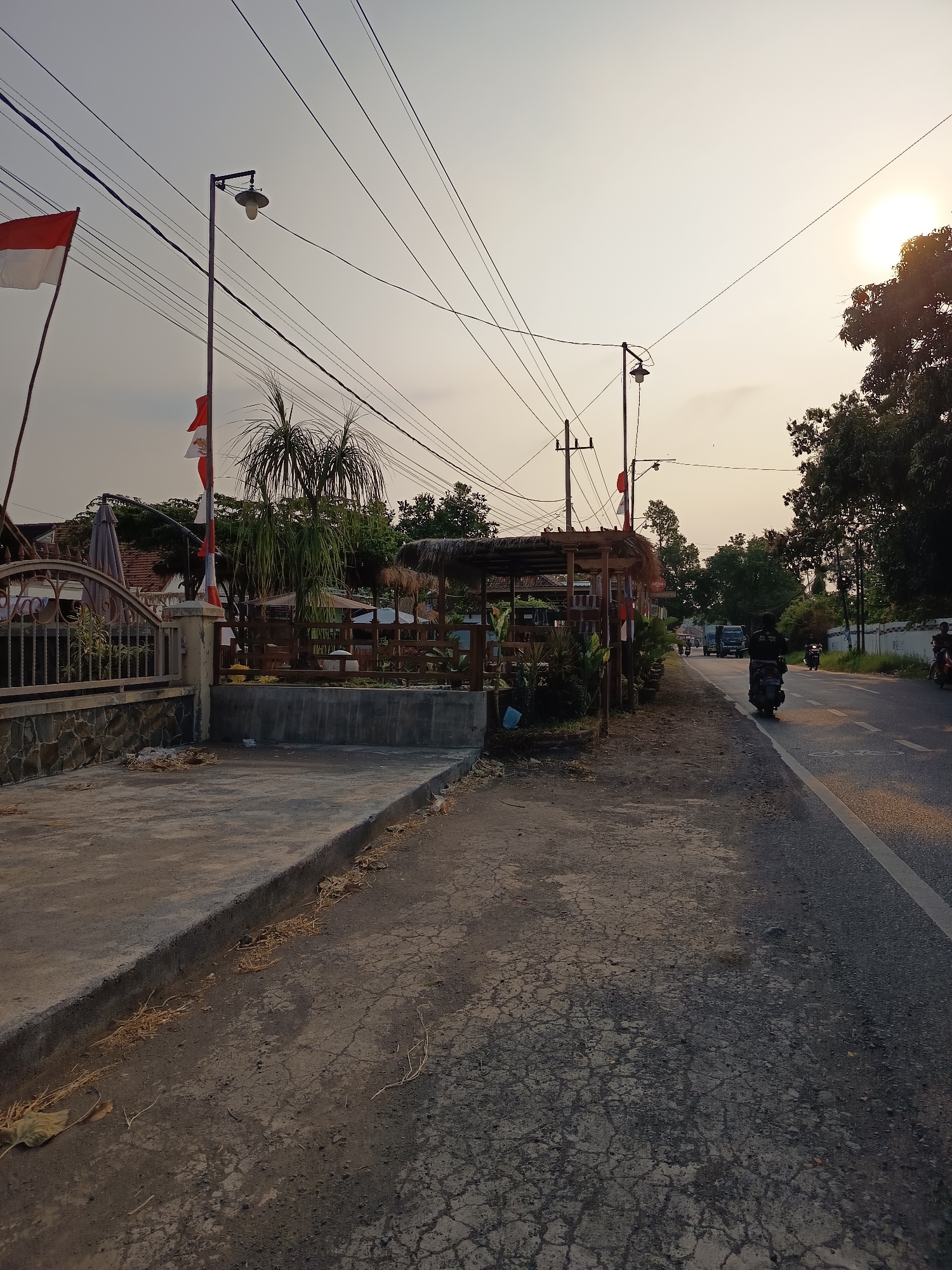 The image depicts a roadside scene at sunset in Kandangjati Wetan, Indonesia. The sun is low in the sky, casting a warm glow. The foreground features a cracked asphalt road and a concrete sidewalk bordered by dry grass and leaves. On the left, a metal fence with stone pillars encloses an area with several thatched-roof gazebos and some greenery, including a palm tree. Indonesian flags are visible, one larger red and white flag flying from a pole, and smaller ones adorning the gazebos. Overhead, a complex network of power lines crisscrosses the sky. Beyond the gazebos, trees and buildings are visible. The road itself has a white line marking its edge. Several vehicles are present, including two motorcycles with riders, one traveling in the same direction as the camera and another approaching from the distance. A truck is also seen in the distance. The overall atmosphere is quiet and typical of a rural or semi-rural area at the end of the day.
