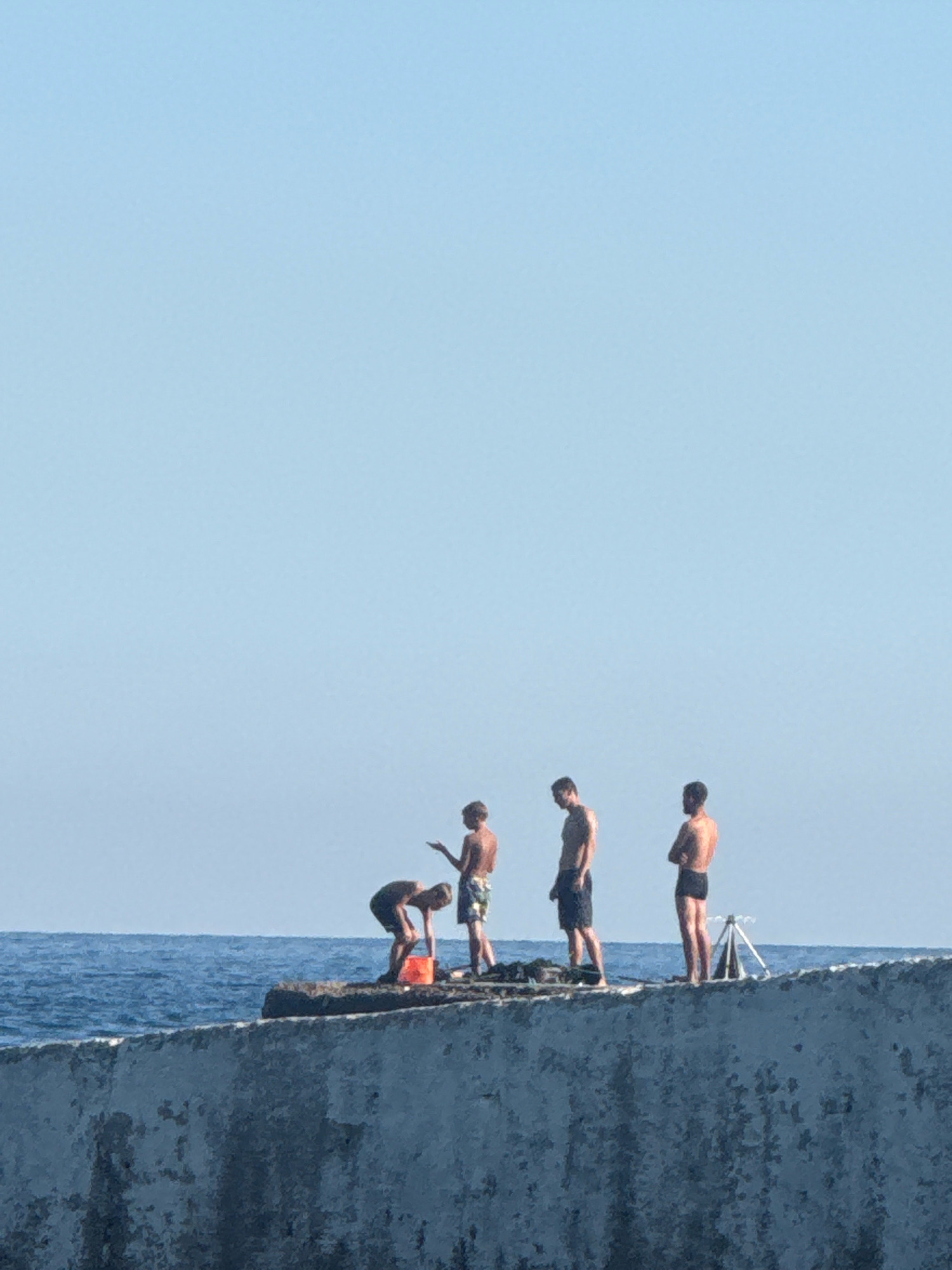 Four individuals are standing on a breakwater at the edge of the sea. The breakwater is constructed from concrete and is textured and weathered. To the left, two younger individuals appear to be engaged in an activity near an orange bucket. One is bent over, and the other is standing and gesturing. To the right, two older individuals are standing, facing different directions. The sea is blue and calm, meeting the sky at the horizon. The sky is a clear, pale blue. There is a small tripod-like object visible on the breakwater to the far right. The scene is likely the coast of Partenit, Ukraine.