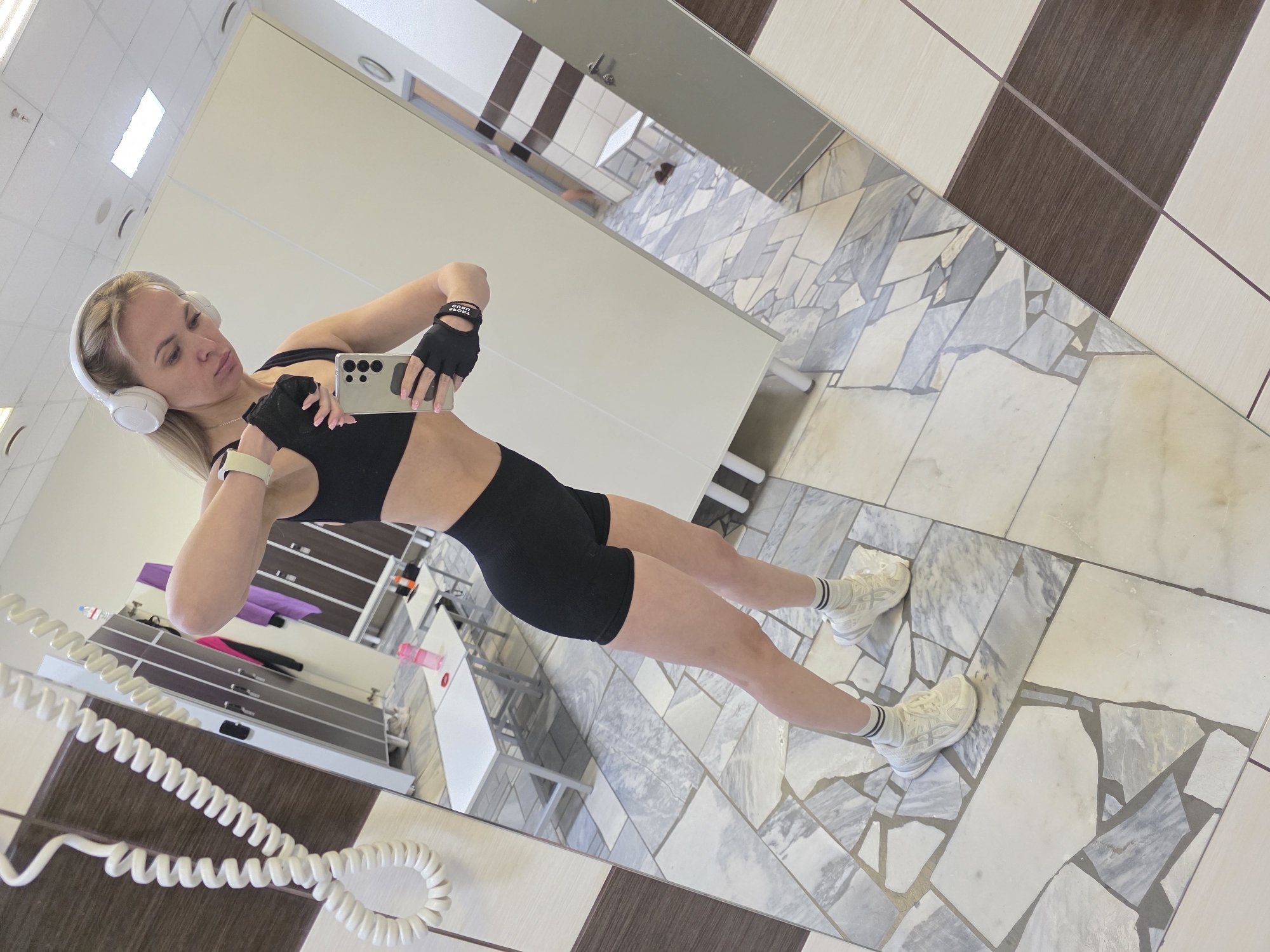 A woman is taking a selfie in front of a mirror in what appears to be a gym or dressing room. The woman is wearing a black sports bra, black shorts, white athletic shoes, and white headphones. She has blonde hair and is wearing a black glove on one hand. She is holding a smartphone in her hand. The background shows a tiled floor, a sink area, and other details suggesting a gym or dressing room setting. The scene is located in the city of Chaikovsky, Russia.