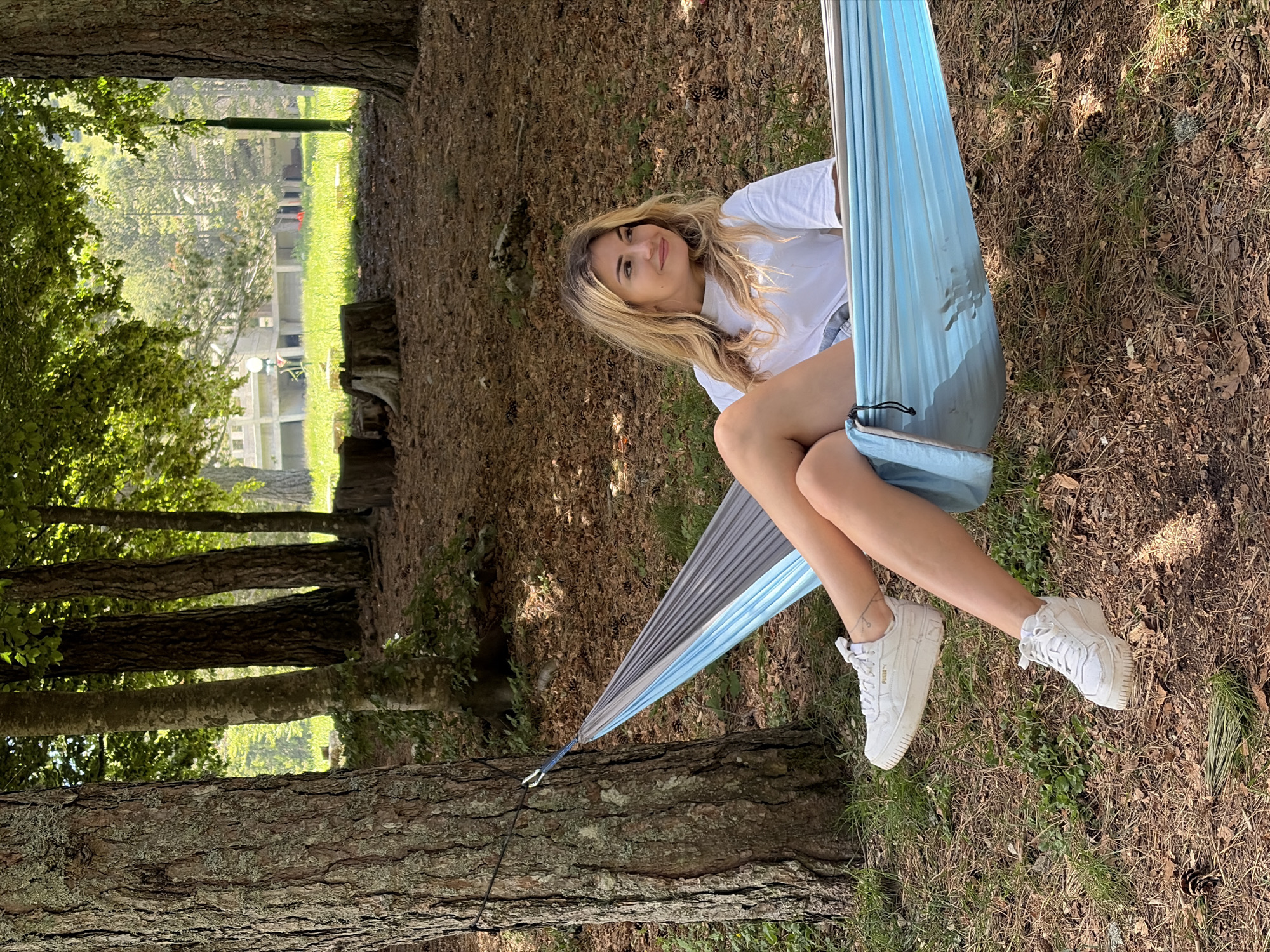 In Mali Bostur, Montenegro, a wide outdoor shot captures a young woman resting in a hammock in a wooded area on a sunny day. The foreground consists of dry leaves, pine needles, and scattered pine cones covering the ground, with interspersed patches of green grass. A multi-colored fabric hammock, primarily light blue and grey, is centrally positioned, strung between two large trees with rough, textured bark.
A light-skinned woman with long, wavy blonde hair is seated in the hammock, looking towards the right with a faint smile. She is wearing a plain white short-sleeved t-shirt and light-colored shorts, with white platform sneakers on her feet, one of which displays a "PUMA" logo on the side. The left side of the hammock is attached to a prominent tree via a black rope and a silver carabiner.
The midground features numerous trees of varying sizes and bark textures, creating a partial canopy with visible green foliage. Sunlight filters through the leaves, casting dappled light on the forest floor. In the background, beyond a grassy clearing, a large, light-colored building with multiple levels and windows is partially visible through the trees, suggesting a residential or institutional structure. Several tree stumps are also visible on the forest floor in the mid-to-background. The overall scene depicts a relaxed moment in a natural, semi-forested environment adjacent to human development.