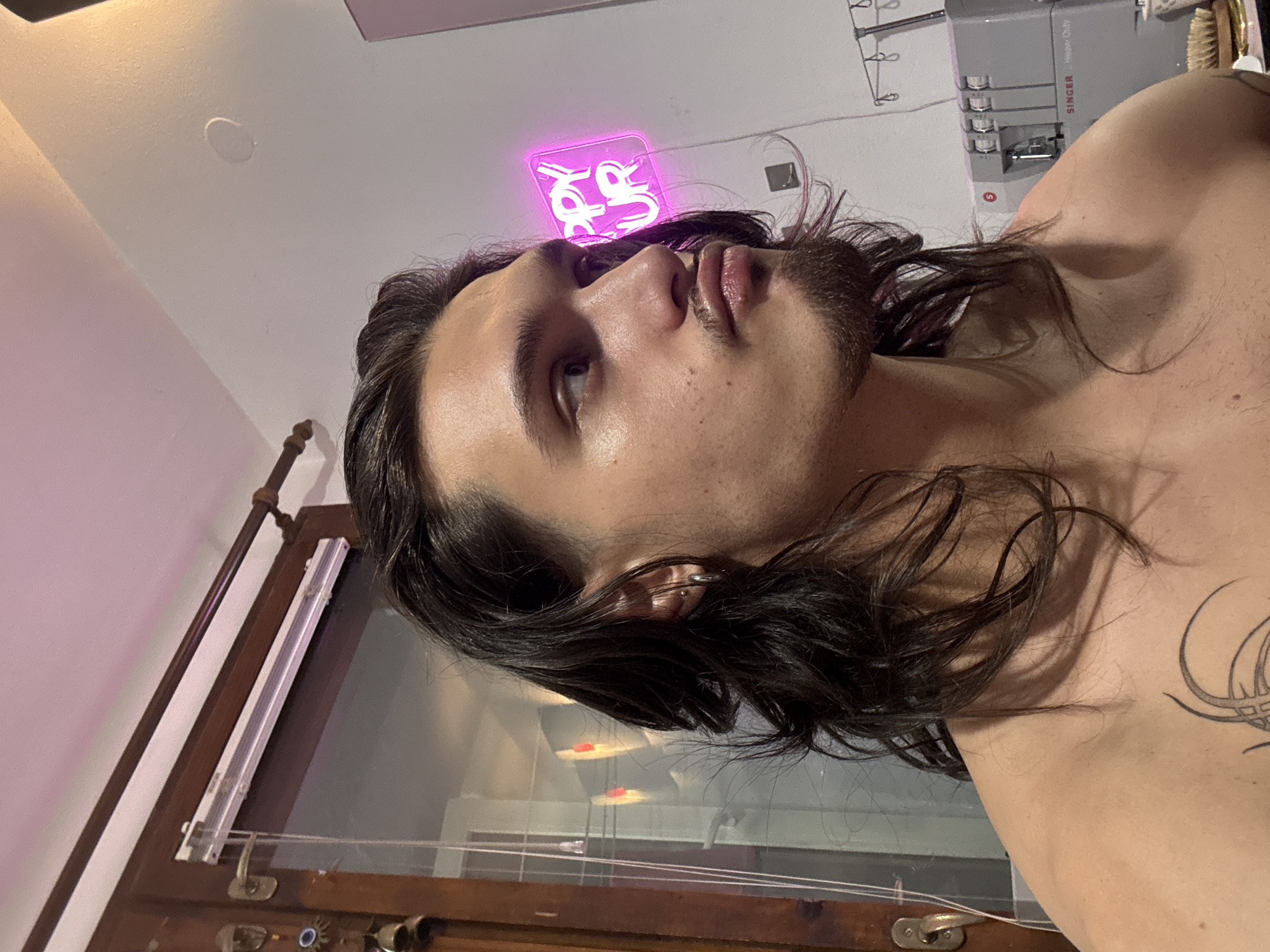 The original image, rotated 90 degrees clockwise, captures a close-up of a person's upper body and face in an indoor setting in Sofia, Bulgaria. The subject is a young individual with long, dark, wavy hair and facial hair (beard/stubble). They appear shirtless, revealing a tattoo with an abstract, curved pattern on their right shoulder/upper arm. A small metallic earring is visible on their left ear. The person is looking upwards and slightly to the left, with their mouth slightly open, conveying a contemplative or neutral expression.
In the background, a bright pink neon sign is visible above the person's head. The sign features stylized text that is difficult to read definitively but appears to be a script or graphic design. Further back, to the right of the subject, a gray "SINGER" brand serger or sewing machine is present. The text "SINGER" and "Happer Overlock" are visible on the machine, along with the distinctive "S" logo. To the left, part of a wooden structure, possibly a window frame or furniture, is seen, with a light fixture or track running along it. A circular overhead light fixture is also visible on the ceiling. The walls are light-colored, suggesting an indoor room. The ambient lighting indicates an artificial light source, likely from the neon sign and other room lights, hinting at an evening or nighttime setting.