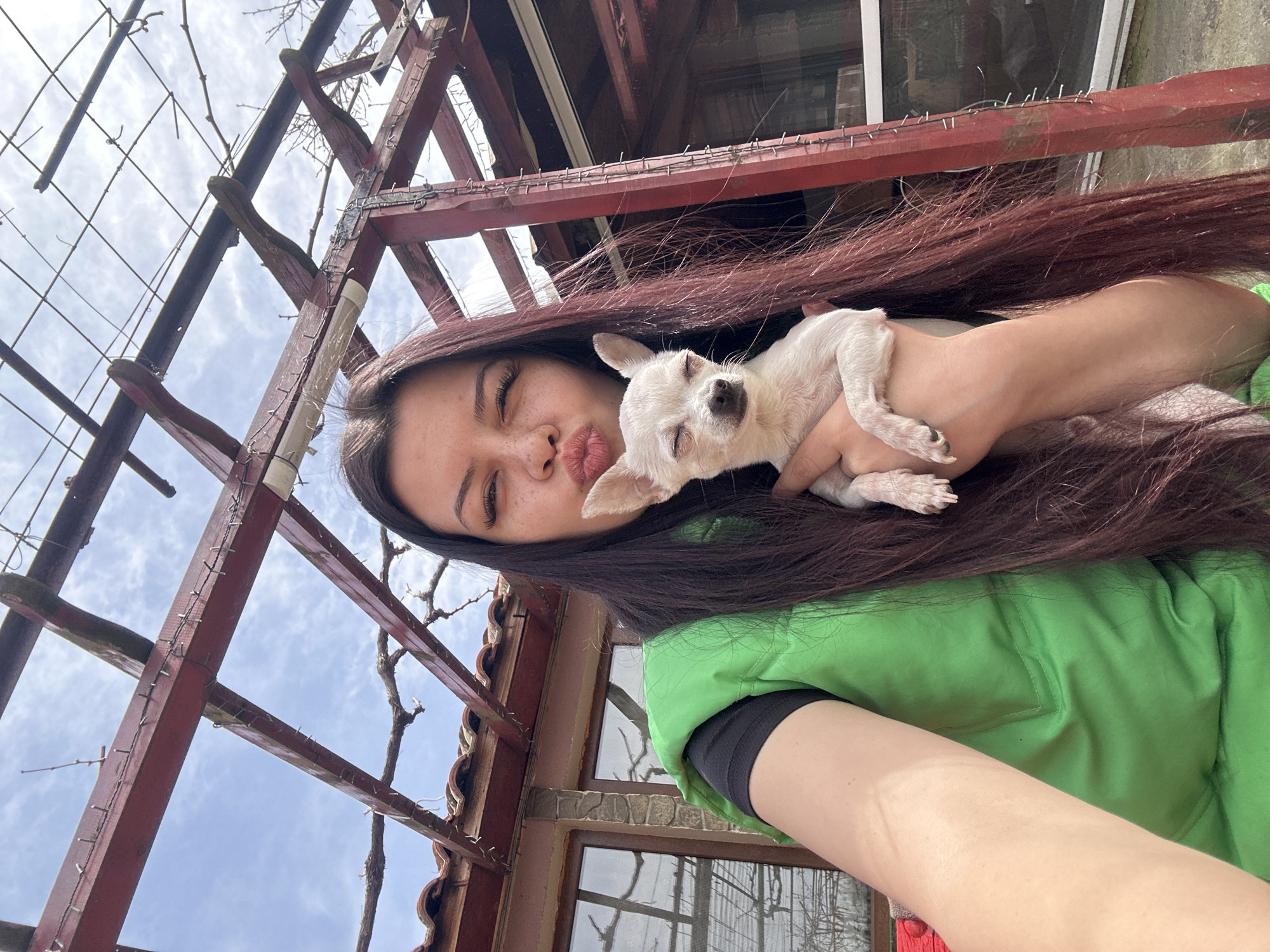 A woman with long brown hair is holding a small white dog in her arms. She is wearing a green shirt with a black underlayer. The woman is making a kissing face. Behind the woman is a wooden trellis structure with sparse vines. In the background, a building with windows is visible. The scene appears to be outdoors, possibly in a yard. Given the city of Chubra, Bulgaria, it could be a residential area.