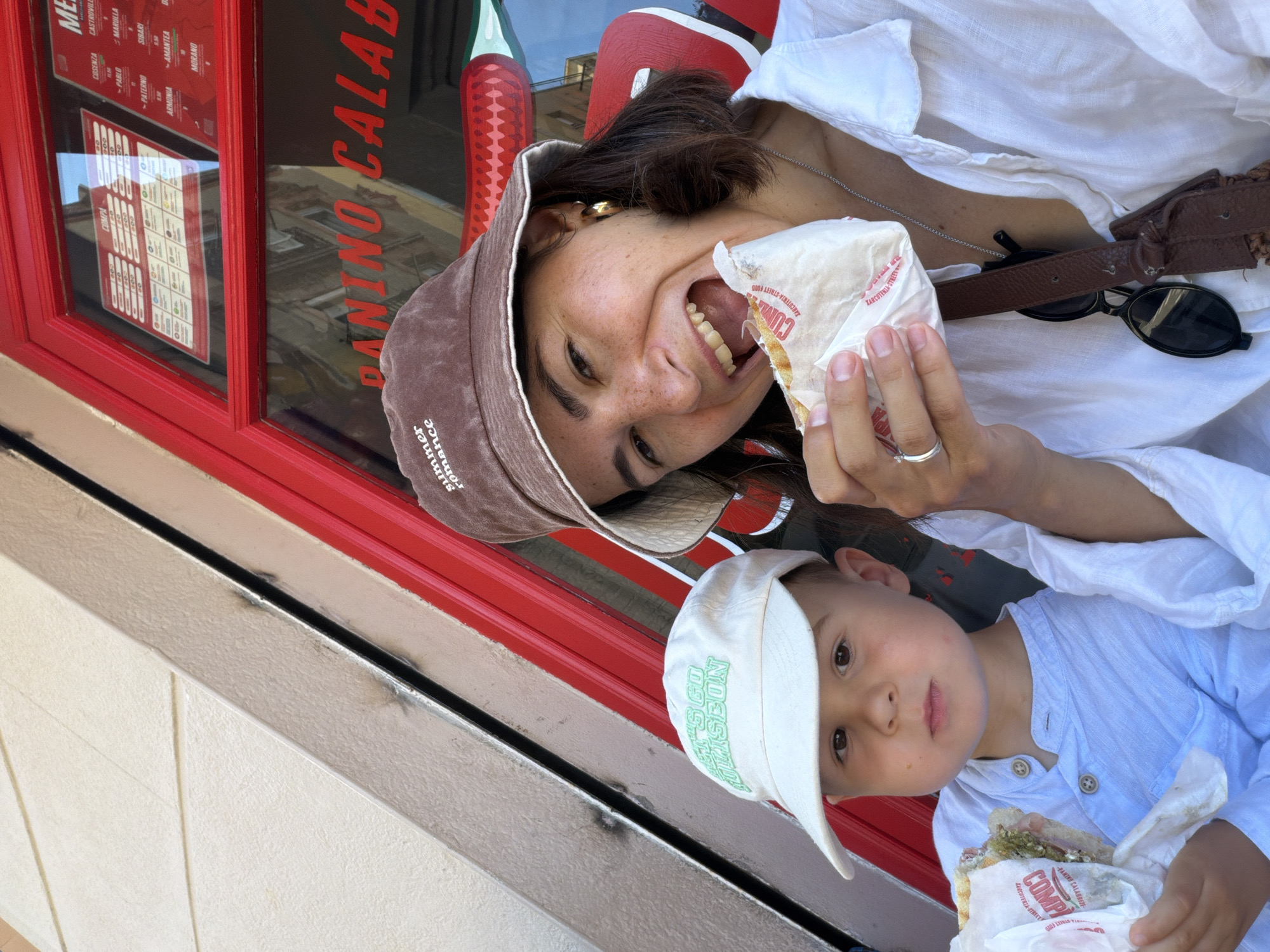 Two people, an adult female and a young boy, are seated outdoors, presumably in front of a shop with a red facade and window displaying menus. The adult female, wearing a brown bucket hat and a white shirt, is smiling and biting into a sandwich wrapped in white paper. The young boy, wearing a white baseball cap and a light blue collared shirt, is holding his own sandwich and looking forward with a neutral expression. The shop's window has red lettering that reads "PANINO CALAB". The overall lighting suggests it is daytime.