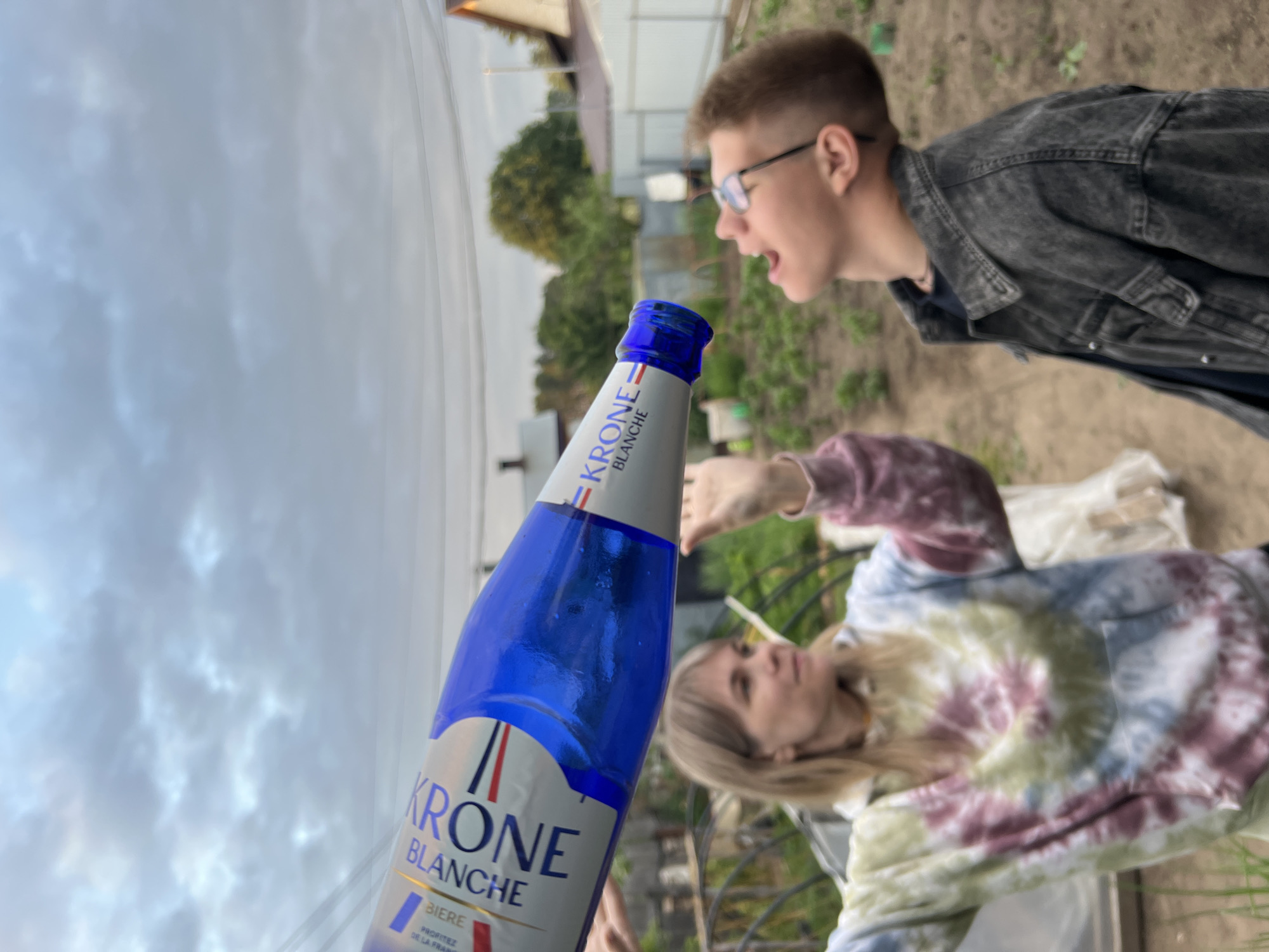 In an outdoor setting that appears to be a backyard garden or dacha in Chaikovsky, Russia, a young person with short, light brown hair and glasses is tilting their head back, mouth open, as if about to drink from a bottle. They are wearing a dark gray denim jacket over a dark shirt. In the foreground, a woman with blonde hair, wearing a tie-dye hoodie in shades of white, yellow, pink, and blue, holds a large white and blue bottle labeled "KRONE BLANCHE BIÈRE" at an angle above the young person. The bottle is the primary focus of the image, positioned diagonally across the frame. The background shows greenery, a fence, and residential buildings typical of a suburban or rural Russian environment. The sky above is overcast with clouds.