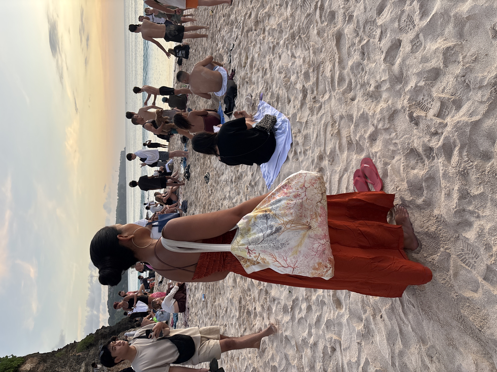 The image depicts a crowded beach at sunset in Kuta Selatan, Indonesia. Many people are present, mostly sitting or standing on the sand. Some are shirtless or in swimwear, suggesting they've been swimming. A woman in the foreground wears a rust-colored dress and a tote bag; she is facing the crowd. Further down the beach, a man in beige shorts is lying down. In the distance, the ocean meets the sky, tinged with colors of the setting sun. The sand is light-colored and shows many footprints.