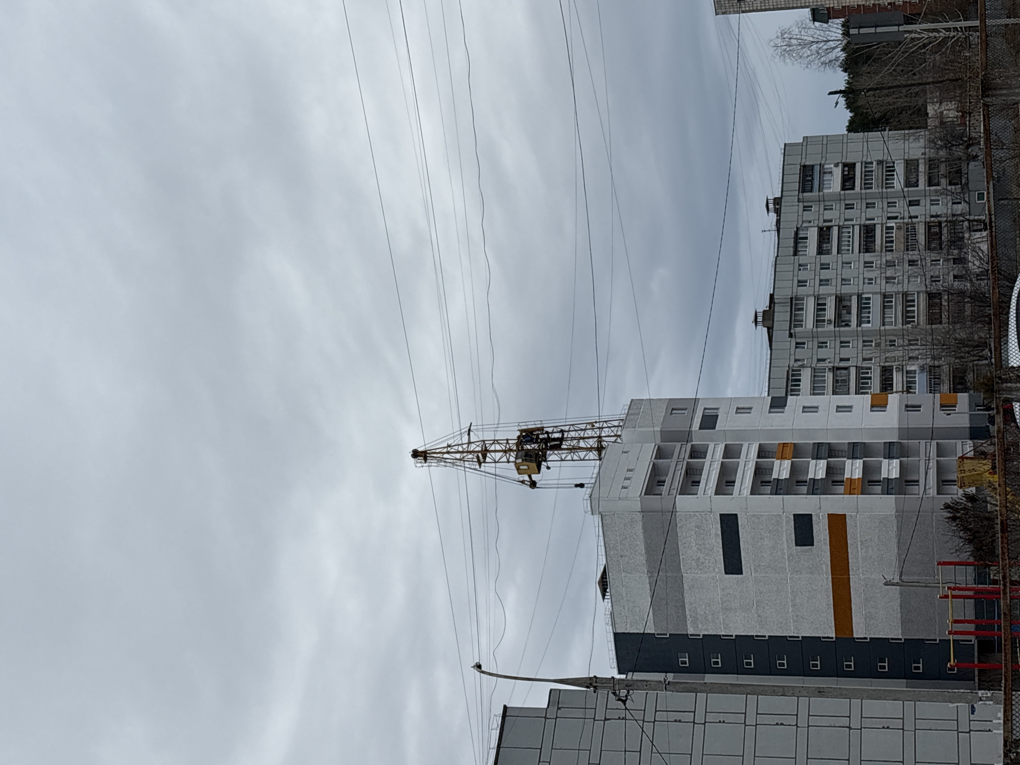 The image depicts a cityscape in Chaikovsky, Russia, under an overcast sky. A construction crane is positioned near several multi-story residential buildings. Electrical wires crisscross the view. The buildings appear to be Soviet-era apartment blocks. A playground is visible in the foreground. No people are visible.