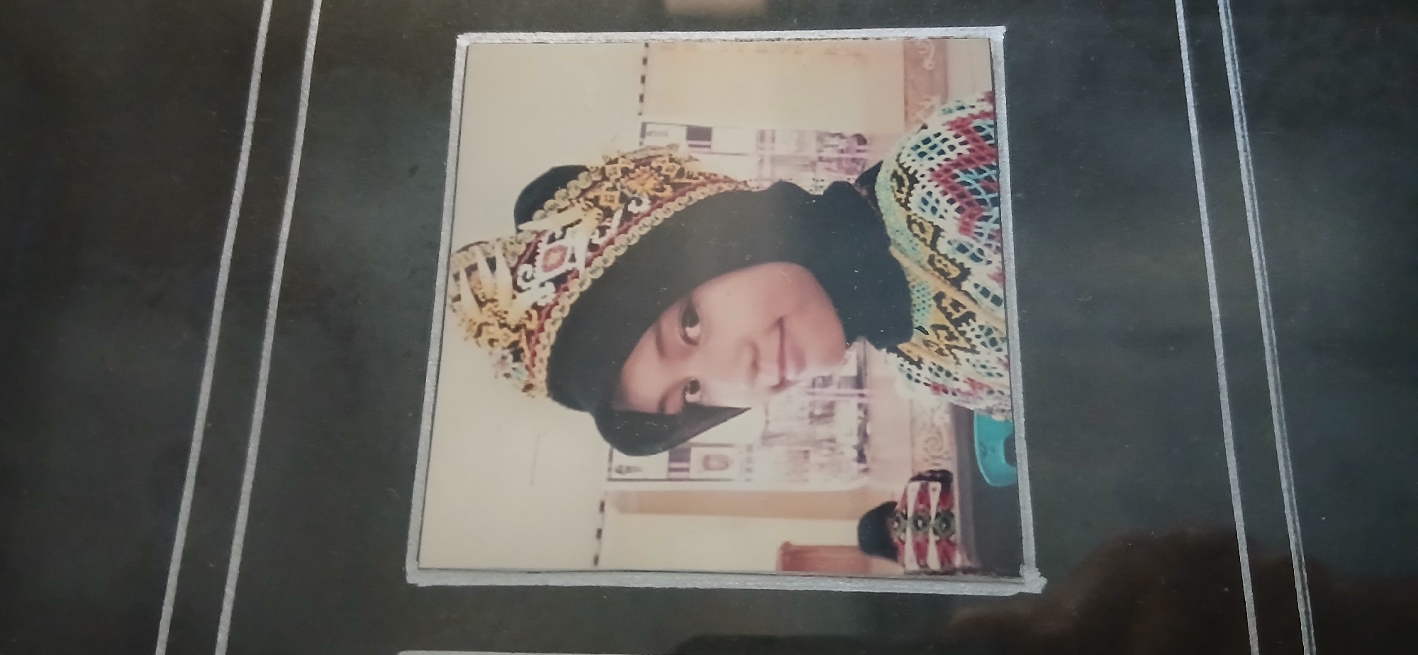 A young woman, likely of Indonesian descent, is depicted in a framed photograph, possibly from a photo album given the dark background with horizontal lines. She is wearing a black hijab and a traditional, ornate headdress with gold, red, and white embroidery. Her attire also includes a colorful, patterned garment around her neck and shoulders, with geometric designs in blue, yellow, red, and white. She is looking towards the right of the frame with a slight smile. The background of the photograph appears to be an indoor setting, possibly a room with light-colored walls and some indistinct objects that could be furniture or decorations. The overall lighting suggests a slightly dim environment. There is no indication of the specific location within Samarinda, Indonesia.