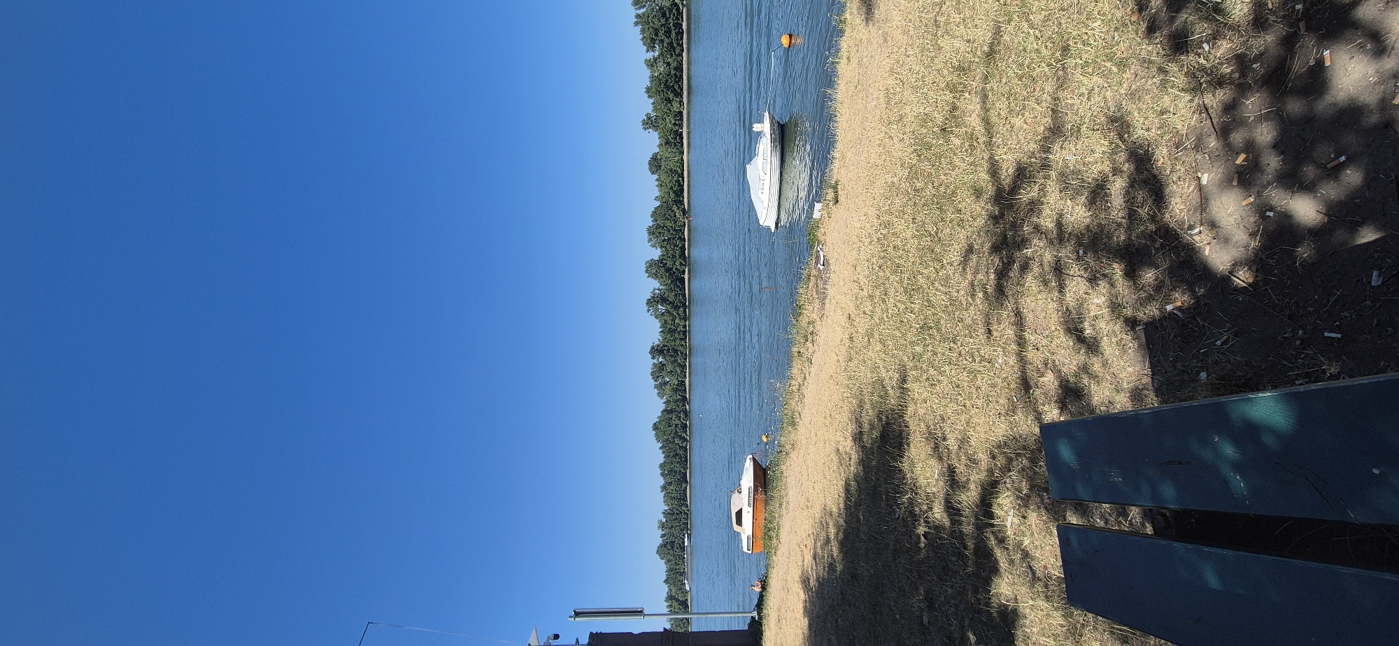 The image depicts a sunny day scene by a body of water, likely a river, with a clear blue sky overhead. In the foreground, a dark green wooden bench is visible, casting dappled shadows from overhead trees onto dry, yellowed grass. The grass is littered with numerous cigarette butts.
On the bank of the water, a small, orange and white boat is moored. Further out in the water, a larger white boat is also moored, covered by a light-colored tarp. The water appears calm and reflects the blue sky. Across the water, a dense line of green trees forms the shoreline of a distant bank, suggesting a park or natural area. A faint, dark pole with a white object at the top is present on the far left of the frame, near the shore.
There is one person visible in the image, sitting on the grass near the orange and white boat on the left bank. No specific actions or interactions are observable. The overall environment suggests a recreational area, possibly a park or a waterfront promenade.
This scene is consistent with a location along the Rhine River in Mainz, Germany, specifically areas like the Rheinuferpromenade or adjacent parks, known for their waterfront recreational spaces and common presence of benches and moored boats.