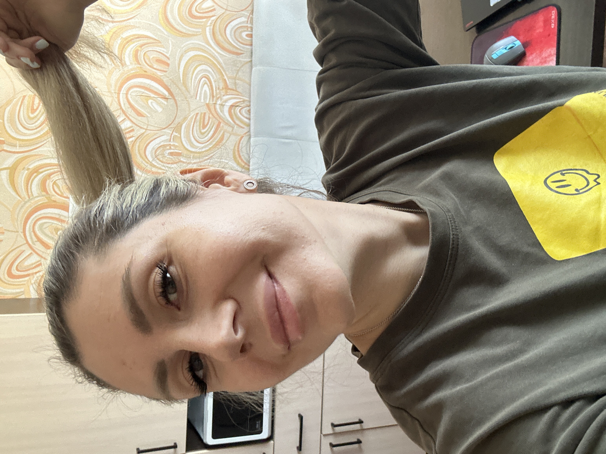 A woman with blonde hair pulled into a ponytail, wearing a green t-shirt with a yellow smiley face graphic, is shown in a close-up portrait. She is smiling and holding a lock of her hair. The background appears to be an indoor setting with light-colored cabinetry, a microwave, and patterned wallpaper. The image is likely taken in Novoaltaysk, Russia.