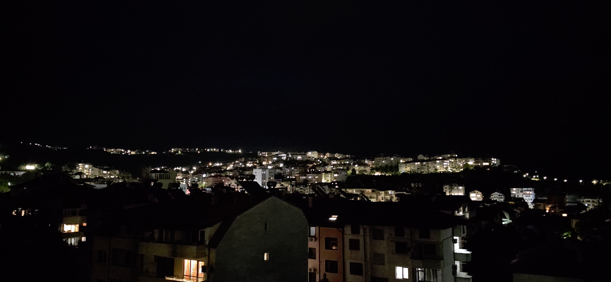 The image presents a wide, high-angle view of a city at night, identified as Sandanski, Bulgaria. The overall scene is dominated by darkness, with a clear, black sky occupying the upper half of the frame, suggesting a calm night with no visible clouds or celestial bodies.
In the foreground, the silhouettes of multiple buildings are visible, appearing as dark masses against the illuminated city beyond. Several of these buildings have lit windows, emitting warm, yellowish light that hints at interior activity. A large, dark building with a single bright window stands prominently in the lower-center of the image.
The midground and background reveal the expansive city, characterized by numerous residential and commercial structures sprawling across hilly terrain. Thousands of electric lights, mostly warm white and yellow, dot the landscape, outlining the urban development and extending far into the distance. These lights create a vibrant, glowing pattern that contrasts sharply with the dark hillsides and the night sky. The distribution of lights suggests the city is built on undulating topography, with buildings and streets ascending and descending slopes. No specific activities or individuals are discernible, and there is no visible text. The scene evokes a sense of peaceful urban life after dark.