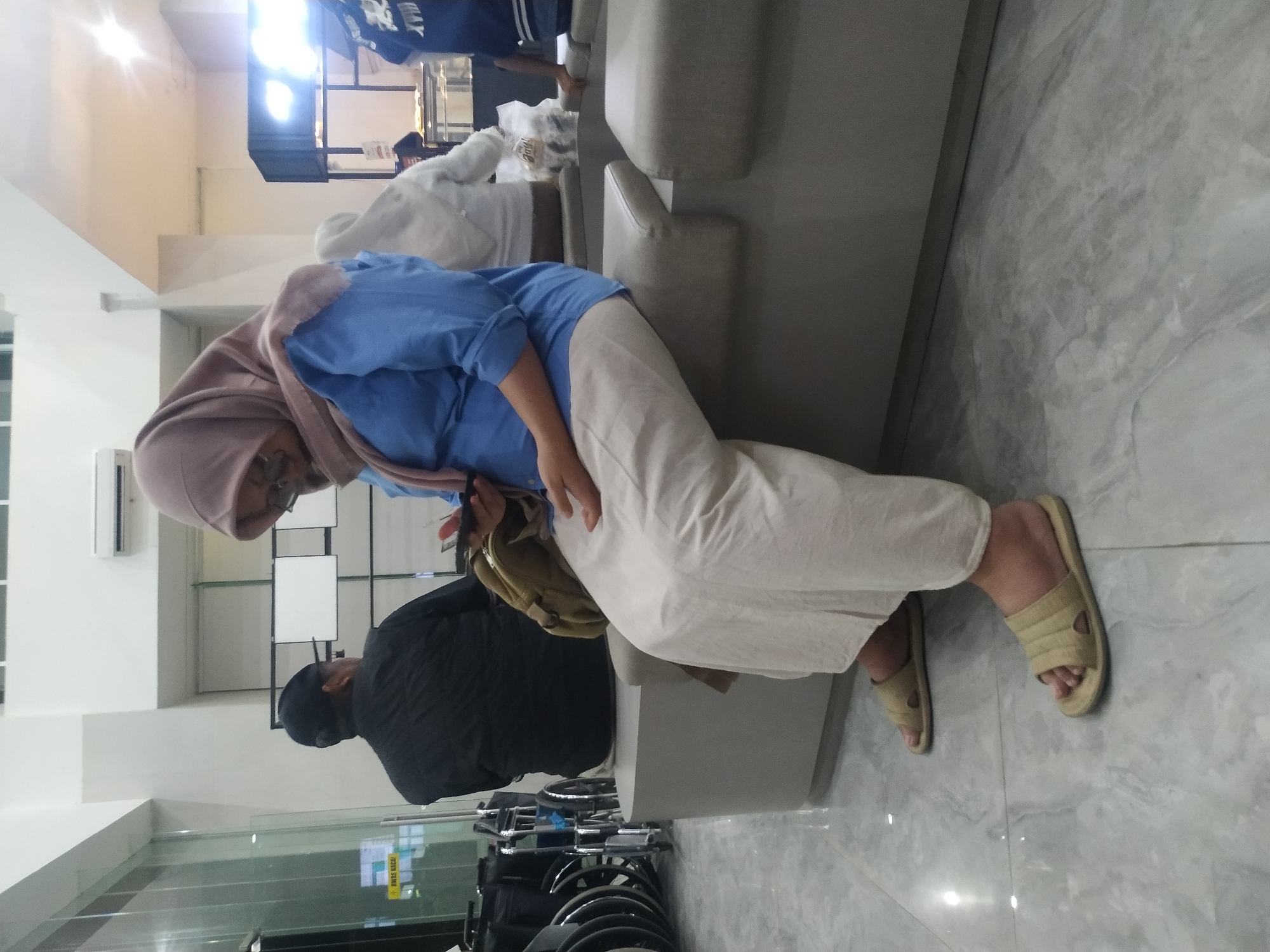 In Mojosongo, Indonesia, a woman wearing a pink hijab and a blue top sits on a gray bench in a well-lit indoor space, possibly a waiting area. She is holding a black smartphone and appears to be looking at it. Her attire includes a light-colored skirt and tan sandals. To her left, a man in a black jacket and cap is seated, facing away from the camera. Several wheelchairs are visible in the background, to the left of the man. The floor is tiled with a gray, marbled pattern. A sign with "AWAS KACA" which translates to "CAUTION GLASS" is visible on a glass panel to the left. The environment suggests a public building, potentially a hospital or clinic.