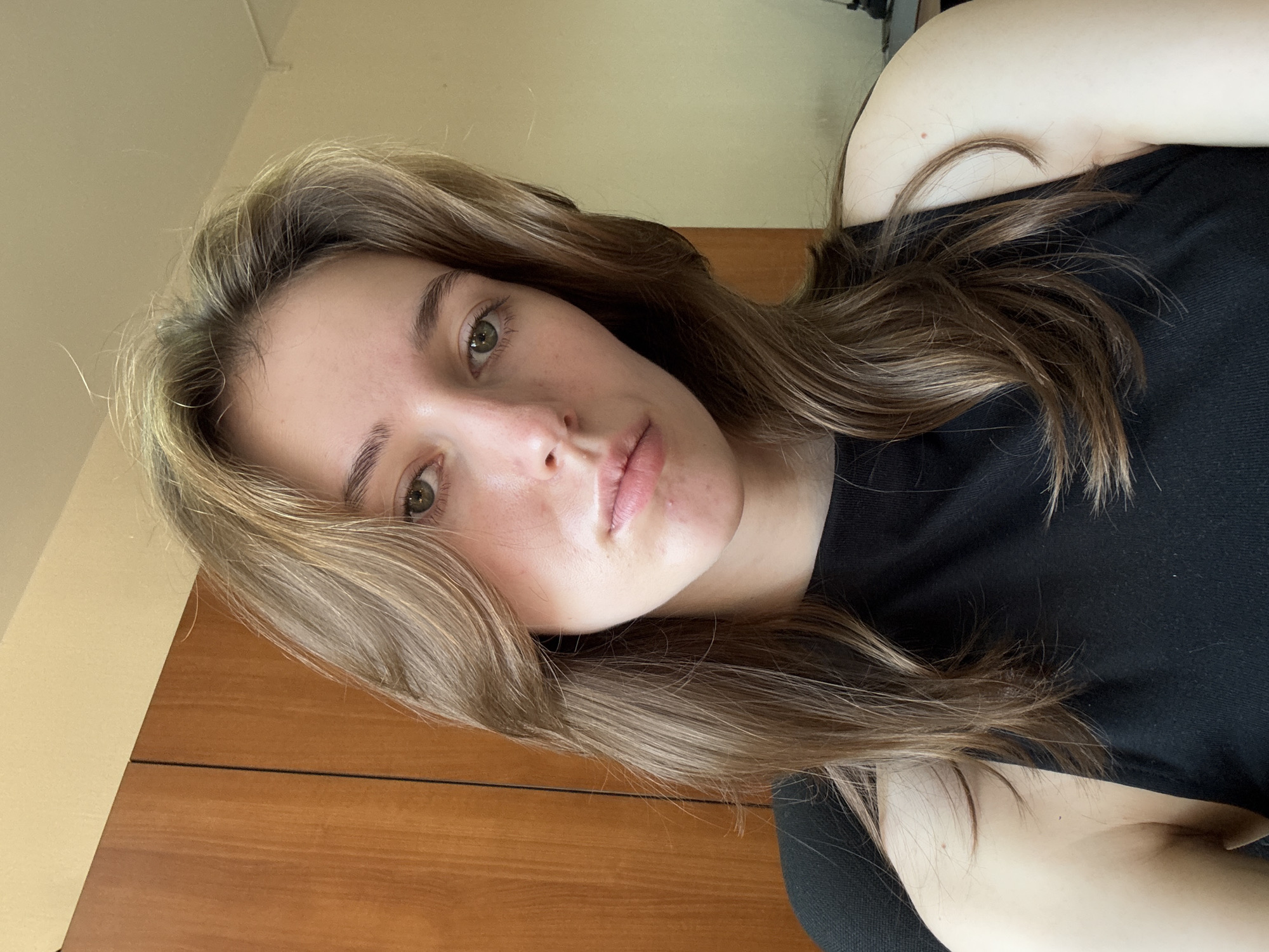 A young woman with long, light brown hair and green eyes is looking forward, her head slightly tilted. She is wearing a black sleeveless top. The background shows a wooden cabinet or door on the left and a plain wall on the right. The lighting suggests an indoor setting. There is no indication of the city of Perm, Russia, or any specific location within a city.