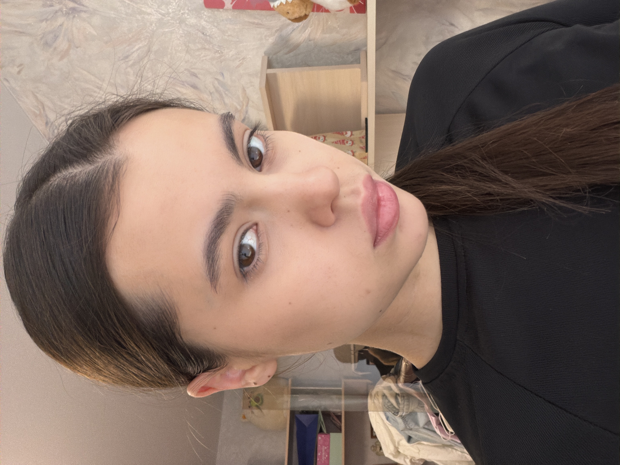 A young woman with brown hair pulled back in a ponytail and wearing a black top is looking directly at the camera. The background includes a shelf, various packages, and a patterned surface. The location is unknown, with no clear indication of Chaikovsky, Russia.
