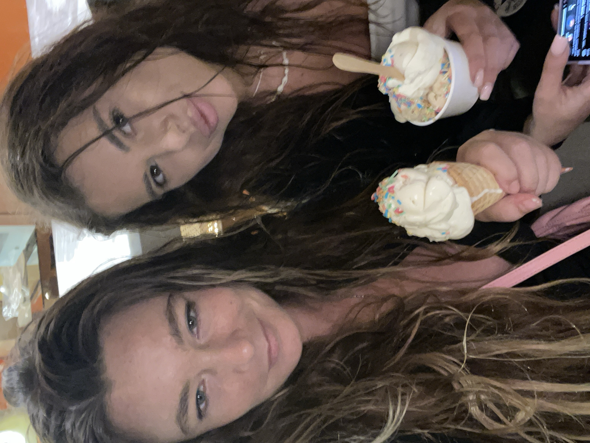 In North Kuta, Indonesia, two women are pictured enjoying ice cream. The woman on the left, with long, light brown hair, smiles as she holds a waffle cone filled with ice cream and colorful sprinkles. To her right, the second woman, with long, dark brown hair, has her mouth slightly open as she looks towards the camera, holding a cup of ice cream with sprinkles and a wooden spoon. Both women appear to be in an indoor setting, possibly a cafe or shop, with a blurred background of warm-toned lights and reflective surfaces.