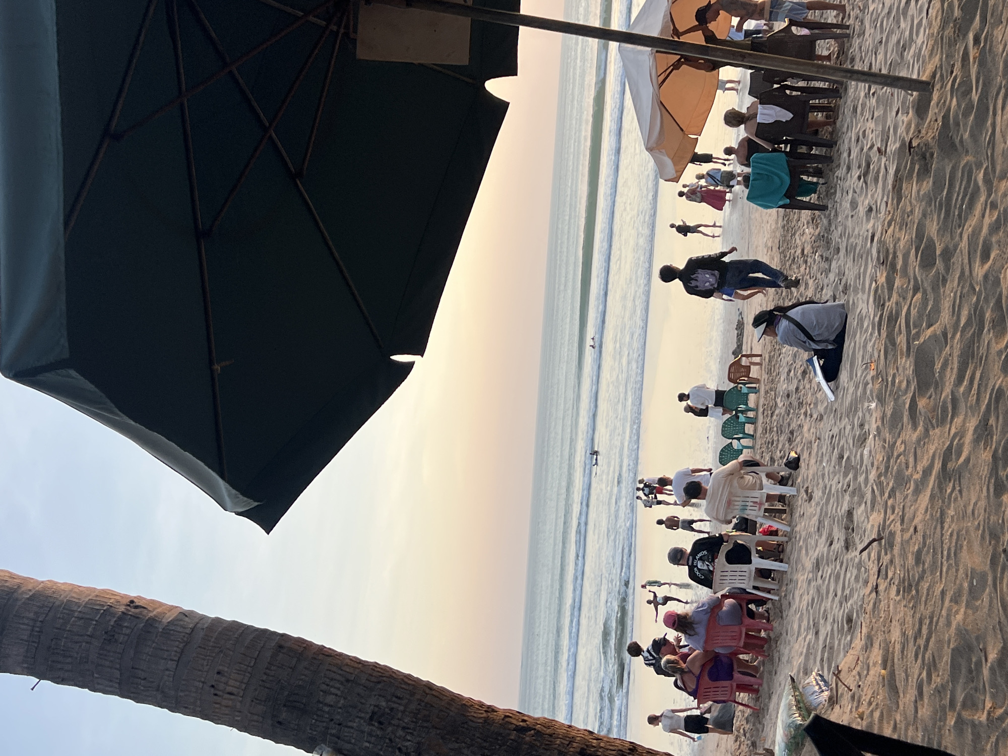 This image captures a beach scene in Kuta, Indonesia, likely during the late afternoon or early evening, as indicated by the soft, warm lighting. In the foreground, a sandy beach is dotted with various individuals seated on chairs and on the sand, some of whom appear to be tourists. A large, dark green umbrella dominates the upper right portion of the frame, providing shade. To the left, the trunk of a palm tree is visible, its rough texture contrasting with the smooth sand.
Further back, people are gathered on the beach, some standing and others seated. The ocean stretches to the horizon, with gentle waves rolling onto the shore. A few individuals can be seen in the water, some possibly surfing on surfboards. The sky displays a gradient of soft colors, transitioning from a pale yellow near the horizon to a light blue. In the mid-ground, there's another lighter-colored umbrella. The overall atmosphere suggests a relaxed beachside setting.