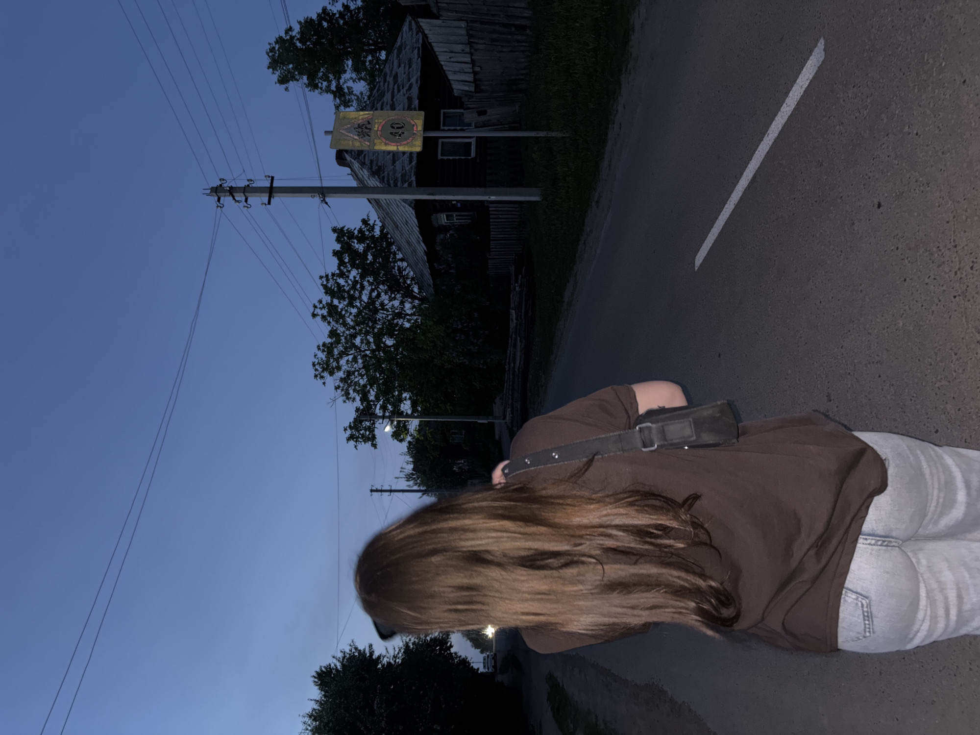 The image depicts a person with long brown hair, wearing a brown shirt and light blue jeans, walking along a paved road. The road has a single white line marking. A wooden building is visible in the background, along with a speed limit sign displaying "40." Utility poles and wires run along the side of the road. The time of day appears to be dusk or evening. The location is Фоки, Russia.