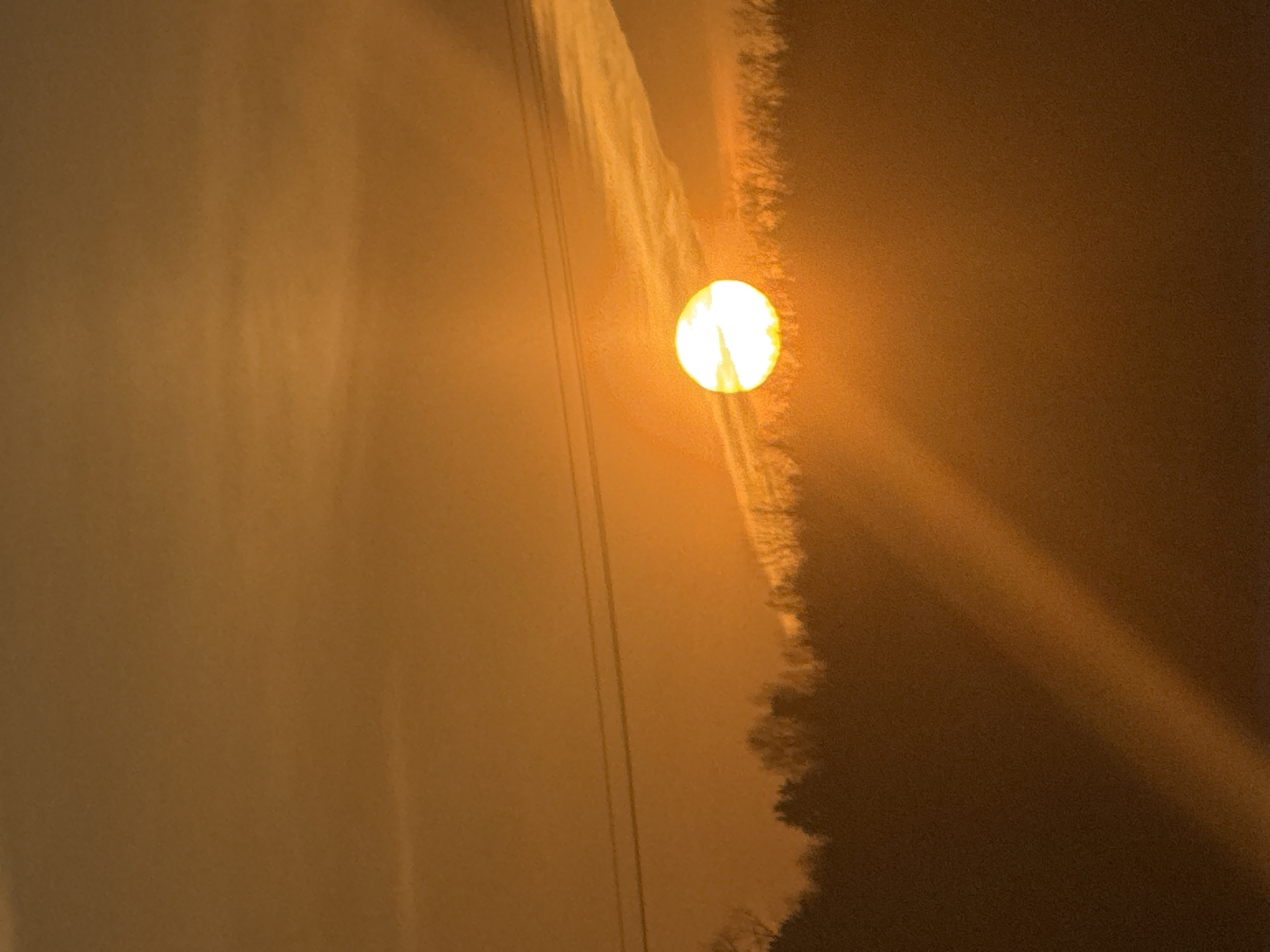 The image shows a sunset in Noviy, Russia. The sun is a bright orb in the center, surrounded by an orange-tinged sky and thin clouds. Power lines are visible on the left side of the image. On the right side of the image, the sun's rays are visible and extend to the right.
