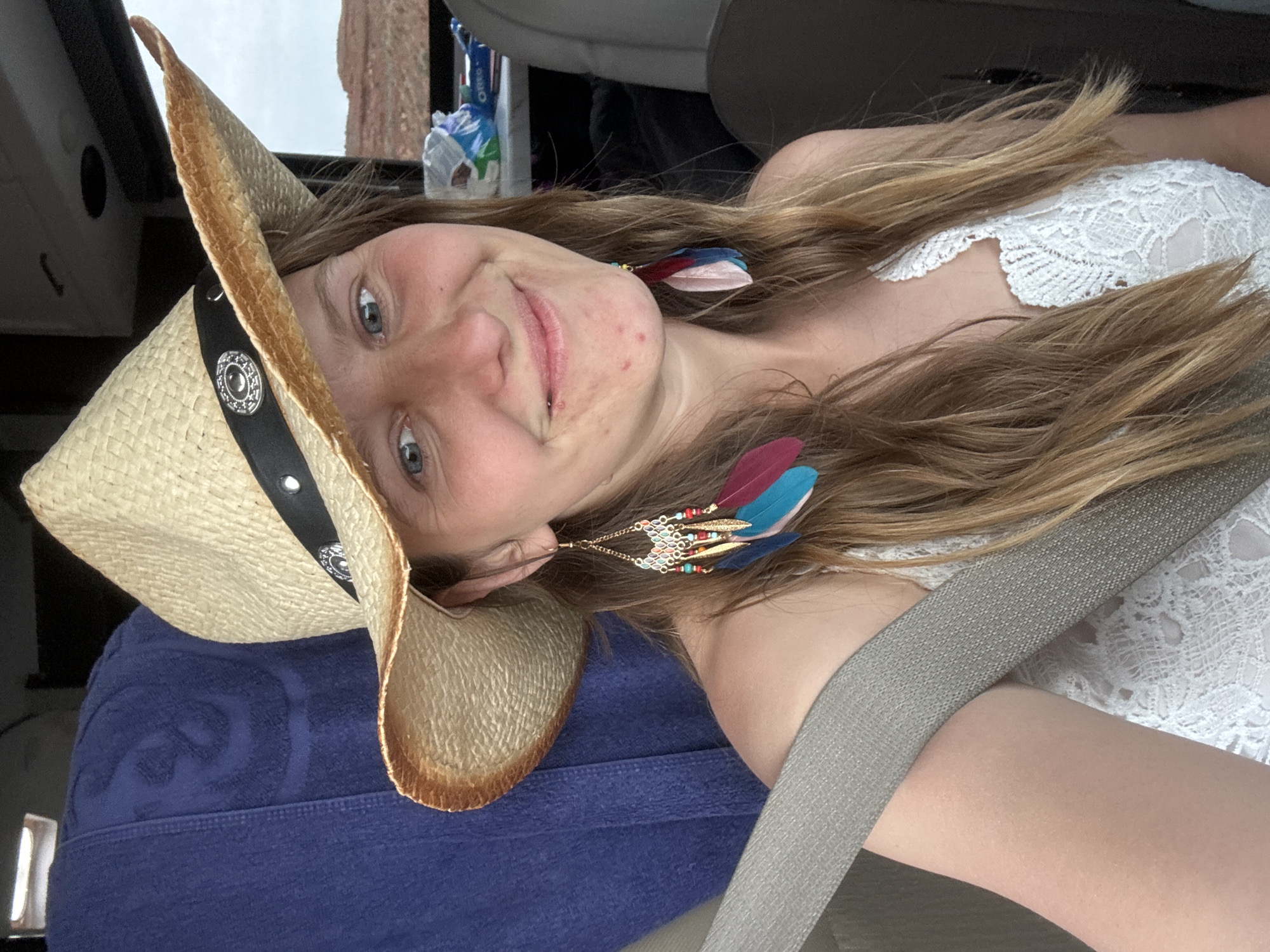 A young woman, wearing a straw cowboy hat, feather earrings, and a white lace top, is shown indoors wearing a gray seatbelt. She is smiling and appears to be in a vehicle, possibly a car. A blue towel and various items are visible in the background. The location is in an unknown area of an unknown city.