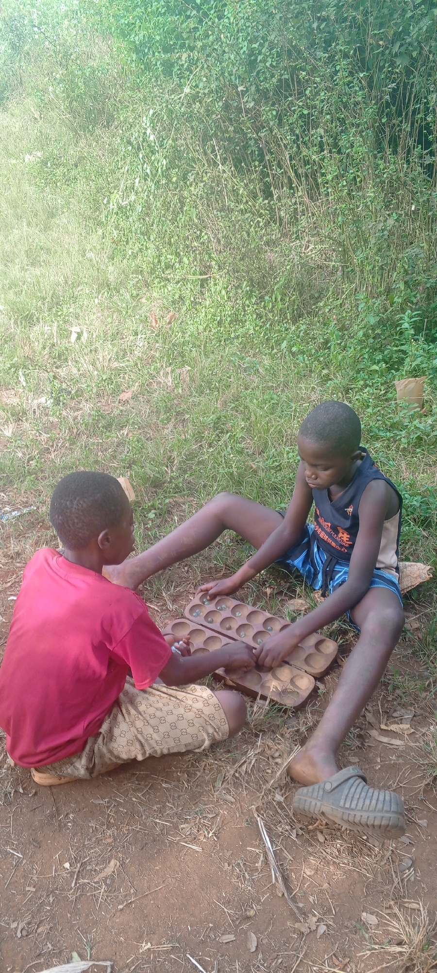 Two children are sitting outdoors on the ground playing a board game with small stones or marbles in the divots. The child on the left wears a red shirt and patterned shorts and is facing slightly away from the camera. The child on the right wears a dark sleeveless shirt with blue striped shorts and a gray clog-like shoe on the right foot, facing toward the game. The game board is between them, filled with small stones. The area around them has dirt and grass and is in a natural, outdoor setting, possibly in Ngalula, Democratic Republic of the Congo.