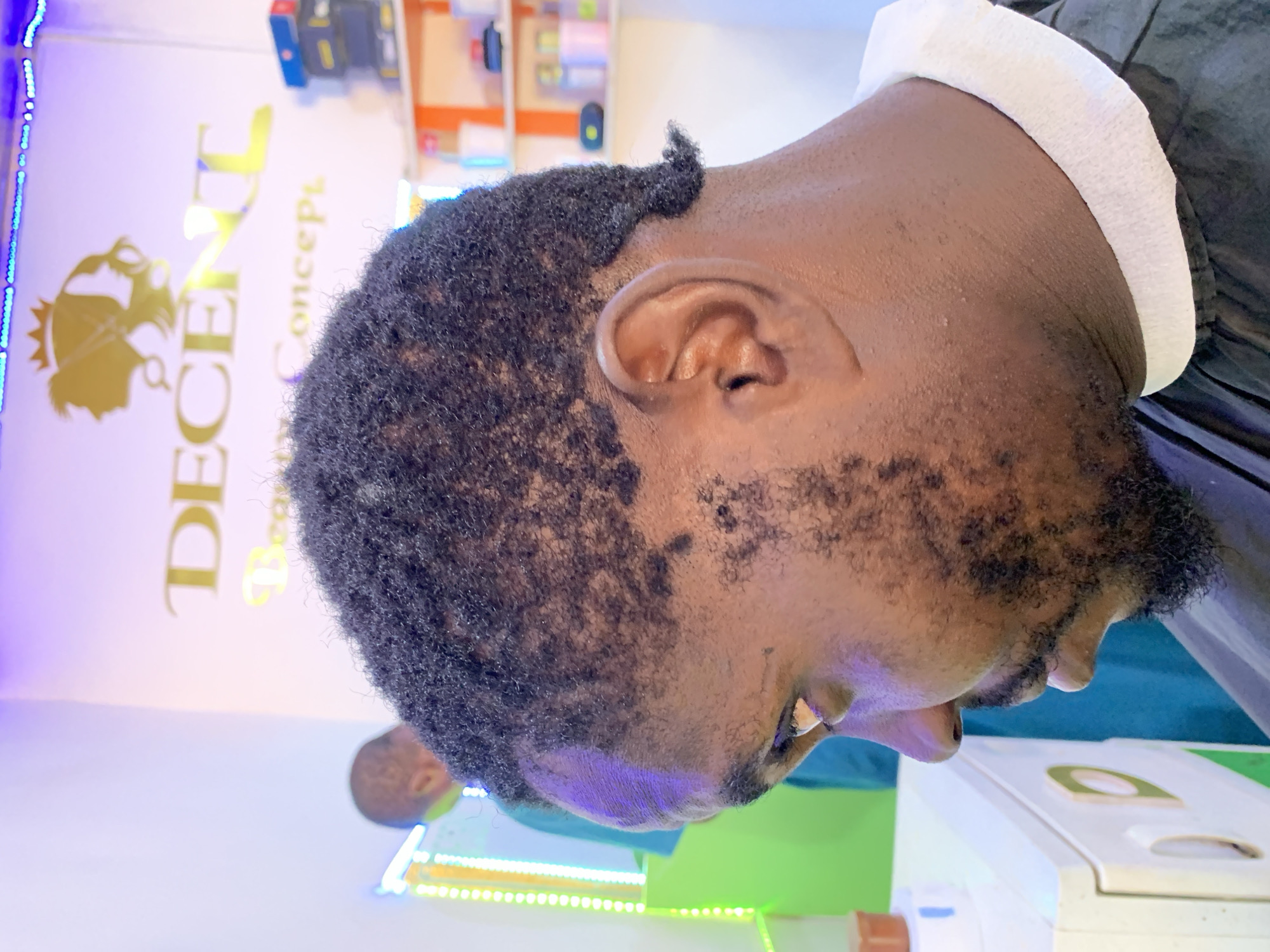 A close-up shot captures the side profile of a man, his dark, short, curly hair and beard prominent. He appears to be receiving a haircut, indicated by the white collar and a dark barber cape. The background is a barbershop, with a sign reading "DECENT Concept" visible on the wall. A shelf with various hair products is partially visible. In the foreground, a green and white machine is present. The location is in the city of Ikorodu, Nigeria.
