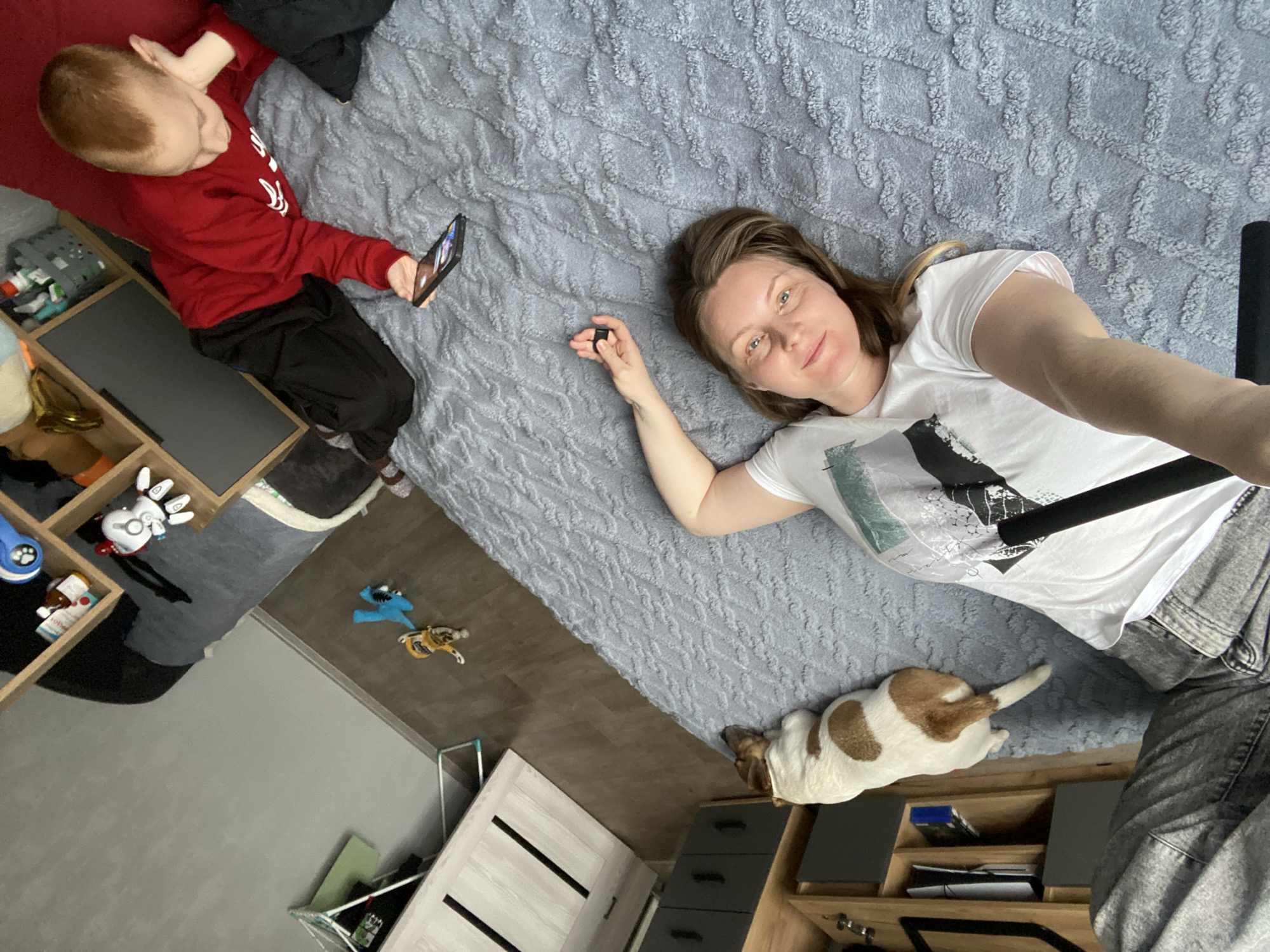 An overhead, rotated view captures a casual indoor scene featuring a woman, a child, and a dog, conveying a relaxed domestic atmosphere.
A woman with light brown hair lies on her back on a light blue, textured blanket covering a bed, positioned in the lower-right of the frame. She wears a white t-shirt with a grey and black abstract design and dark grey pants. Her left arm is bent, holding a small black rectangular object. She smiles subtly, looking towards the camera.
In the upper-left, a young child with short, reddish-brown hair rests on his side on the same blanket, propped against a red cushion. He is dressed in a red long-sleeved shirt and black pants, engrossed in a black smartphone held with both hands.
A small dog, white with brown patches, lies on the dark brown floor next to the bed, near the woman's legs, appearing to rest or sleep.
The room features a dark brown wooden floor, transitioning to a lighter grey carpeted area. To the left of the child, a light wooden shelving unit displays various items including a grey basket, a blue toy, a golden-colored object, a white robot-like toy, and small boxes. On the floor in the mid-left are a blue monster-like toy and a yellow/orange animal figure. Another wooden shelving unit with dark drawers is visible to the right, containing books and other items. A white door with black handles is partially visible in the bottom-left. The scene is brightly lit, suggesting daytime. No discernible text is visible.