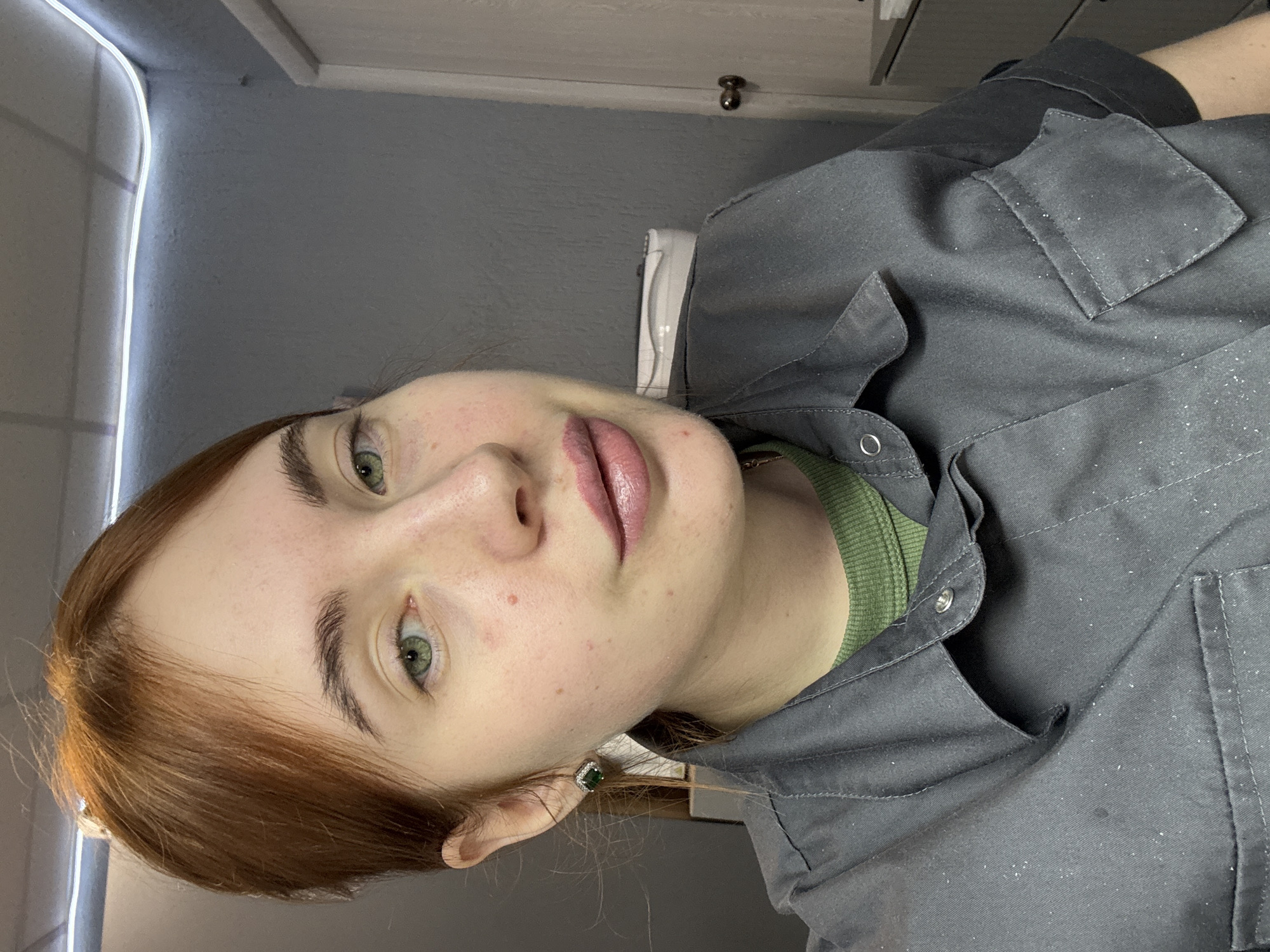 A close-up, slightly rotated, indoor portrait shot of a fair-skinned woman with red hair and green eyes. She is wearing a grey, button-down shirt with a green undershirt visible at the collar. The shirt has a pocket with stitching and appears to be covered in small, white particles. She is wearing a green and silver earring. The background is a grey wall, with a white shelf and light fixture in the upper left. The location is not apparent from the image.