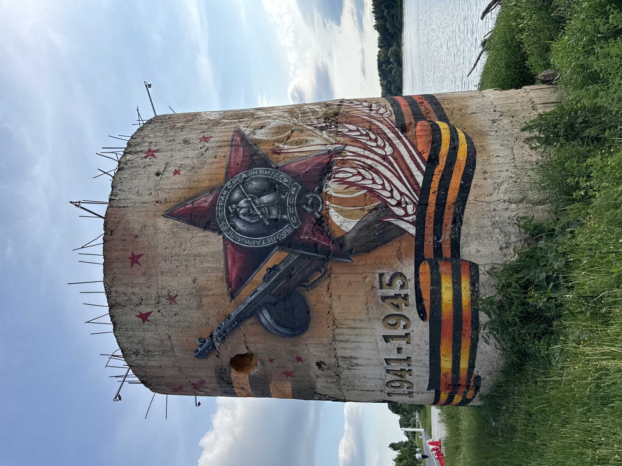 The image shows a cylindrical concrete structure with graffiti depicting a military theme. The graffiti includes a red star with a medal in the center, a machine gun, and the dates "1941-1945". A ribbon with black and orange stripes, resembling the St. George's ribbon, is painted around the base of the structure. Red stars are also painted on the upper part of the cylinder. The structure appears to be unfinished or in disrepair, with rebar protruding from the top.
The structure is located in the city of Kirishi, Russia, near a body of water, possibly a river, and a grassy area with some vegetation. In the background, a wooded area can be seen across the water. There are a few people walking on a paved path to the left of the structure. The sky is partly cloudy.