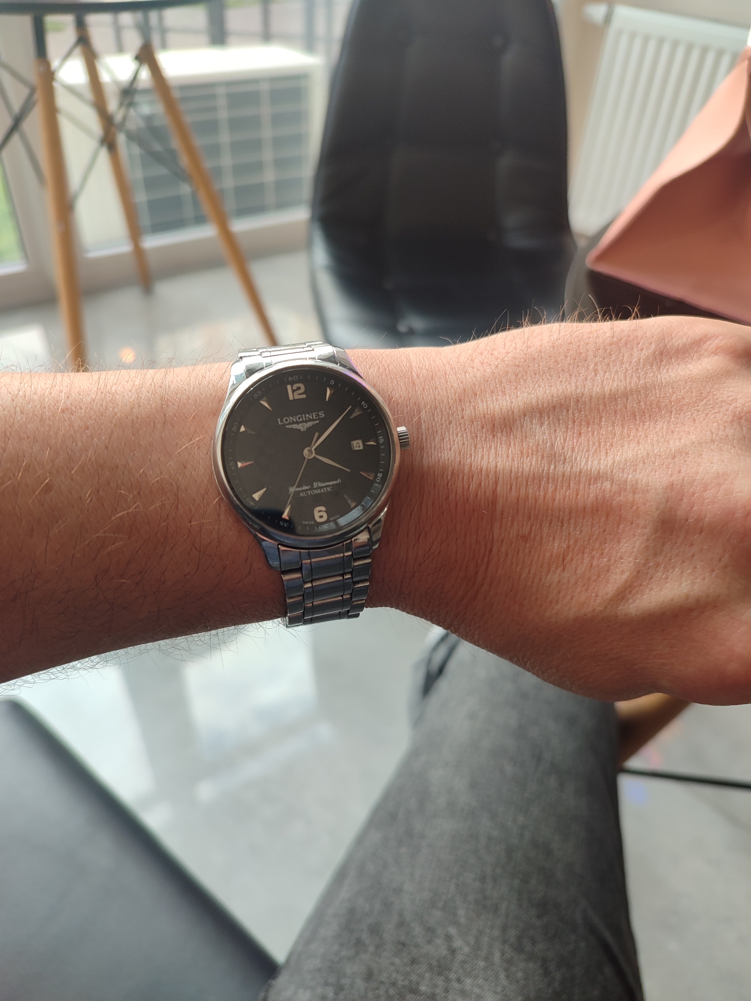 A person's left wrist is shown wearing a Longines automatic watch with a black dial and silver band. The watch has hour markers and a date window. The background is blurred, but appears to be an indoor setting with furniture and windows. The overall lighting suggests an indoor environment during the day.
