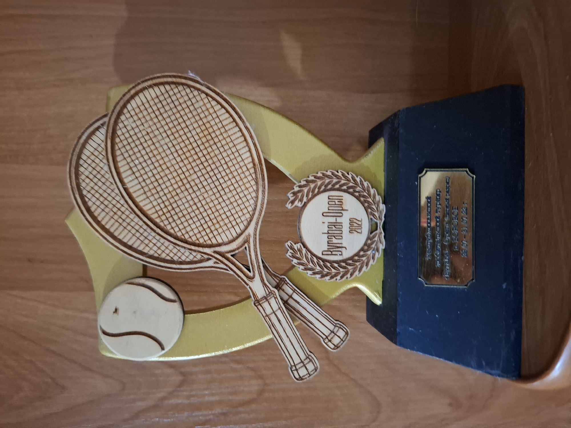 A wooden trophy is displayed on a shelf. The trophy features a gold shield with two wooden tennis rackets and a wooden tennis ball. A circular emblem with a laurel wreath surrounds the text "Byrabai-Open 2022". A gold plaque is affixed to the dark blue base of the trophy, which bears text in Cyrillic script, including "Республиканский турнир" and "г. БОРОБОВЕ 29.07-31.07.22 г." The trophy is positioned on a wooden shelf in what appears to be an indoor setting, with a wooden paneled wall in the background. The plaque indicates that the event took place in Borovoe, Kazakhstan, and the text mentions "Republican tournament". Lisakovsk is a city in Kostanay Region, Kazakhstan, and while the plaque references Borovoe, which is also in Kazakhstan, there is no explicit indication of the trophy's location within the city of Lisakovsk itself.