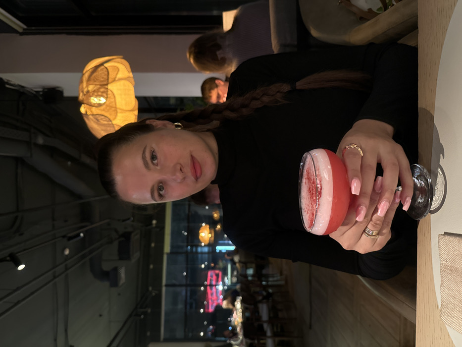 A woman with braided hair holds a pink cocktail in her right hand, adorned with rings and light pink nails. She wears a black long-sleeved top. In the background, a restaurant setting is visible with hanging lights and blurred figures. The image is taken at an angle, with a glimpse of the restaurant's interior.