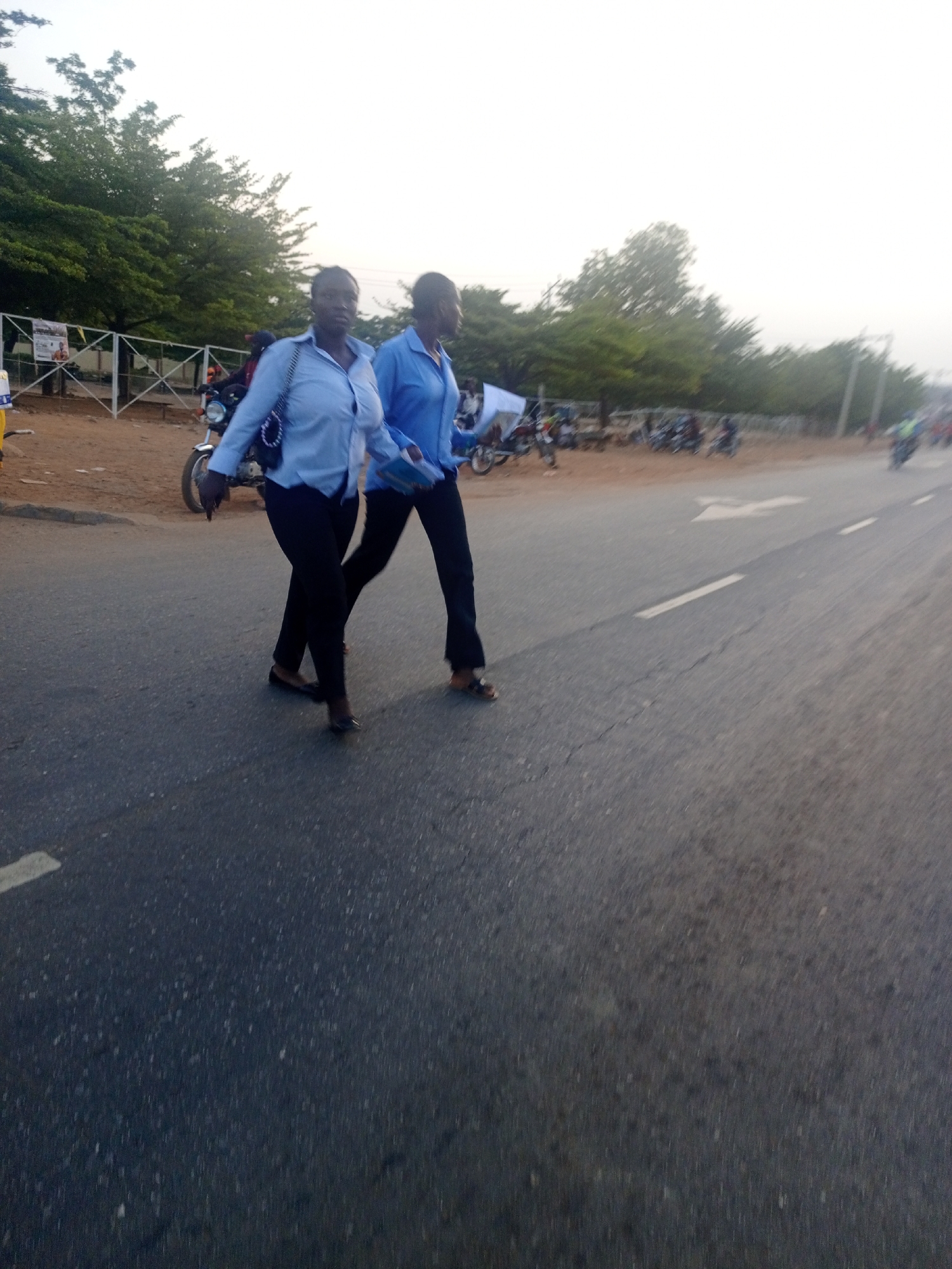 The image shows two women walking along the edge of an asphalt road in Keffi, Nigeria, likely during the late afternoon or early evening. Both women are dressed in light blue long-sleeved shirts and dark trousers, and are walking towards the right side of the frame. The woman on the left carries a dark shoulder bag, while the woman on the right holds papers or a thin book.
The road features a white dashed line and a white arrow painted on its surface, indicating traffic flow. To the left of the walking women, there is a dirt shoulder where several motorcycles are parked. A white metal fence with a criss-cross pattern runs behind the parked motorcycles, and lush green trees line the background. Further down the road, more blurred motorcycles and indistinct structures are visible, suggesting a busy thoroughfare. The sky is bright and diffused, indicating clear weather. A poster with red and yellow accents is attached to the fence in the background, but its text is illegible due to blurriness. The scene captures an everyday moment in a semi-urban Nigerian setting.