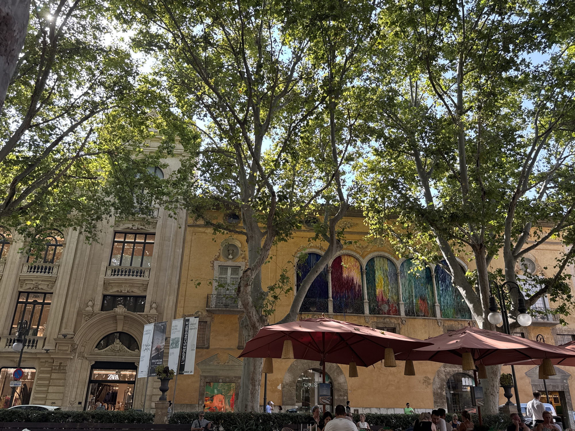 The image shows a view looking up at trees with dappled sunlight filtering through their leaves, in front of buildings in Palma de Mallorca, Spain. The buildings appear to be a mix of architectural styles, with ornate detailing on the facade of the lighter-colored building on the left, and a vibrant, colorful tapestry or mural displayed across the arched windows of the yellow building on the right. Several people are visible sitting at tables under red umbrellas in an outdoor seating area in the foreground, suggesting a cafe or restaurant. Some people are also walking or standing in the background. Banners advertising events or businesses are displayed outside the buildings. The overall impression is of a lively street scene on a sunny day.