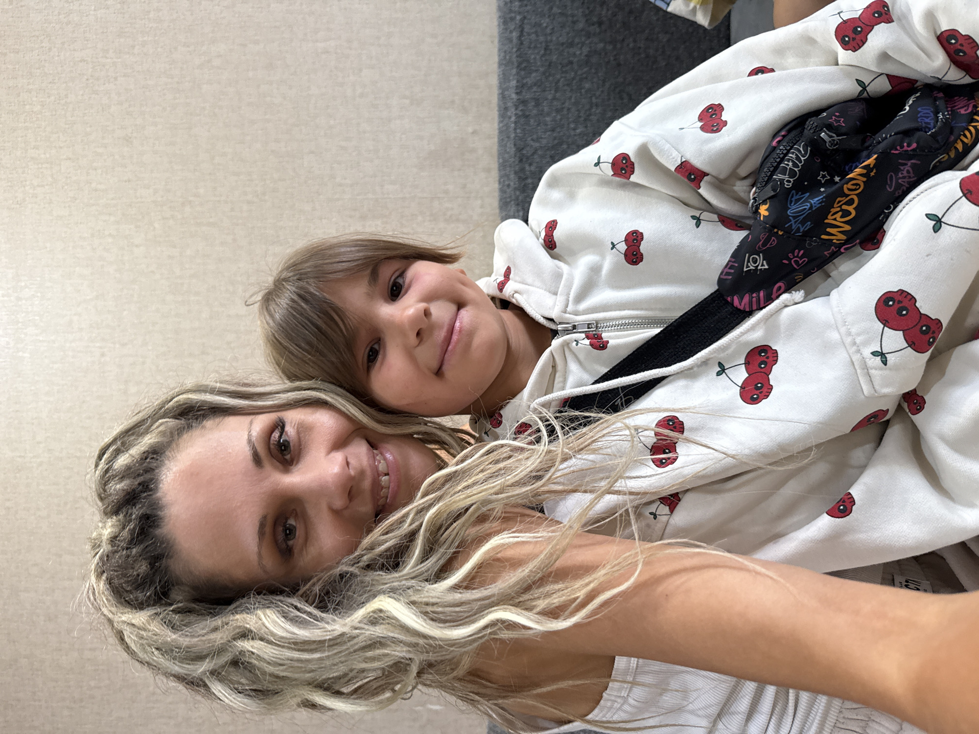 A selfie is captured from a close-up perspective, featuring two individuals, likely mother and child, seated on a couch. The woman on the left has blonde, wavy dreadlocks and is wearing a white tank top. She is positioned in the foreground, with her arm around the child. The child on the right has brown hair and is wearing a white zip-up hoodie adorned with a cherry skull print. A black bag with colorful graffiti is slung across the child's chest. The background consists of a textured, light-colored wall and a dark gray couch. The image does not contain any explicit information about the location being Rusanovo, Russia.