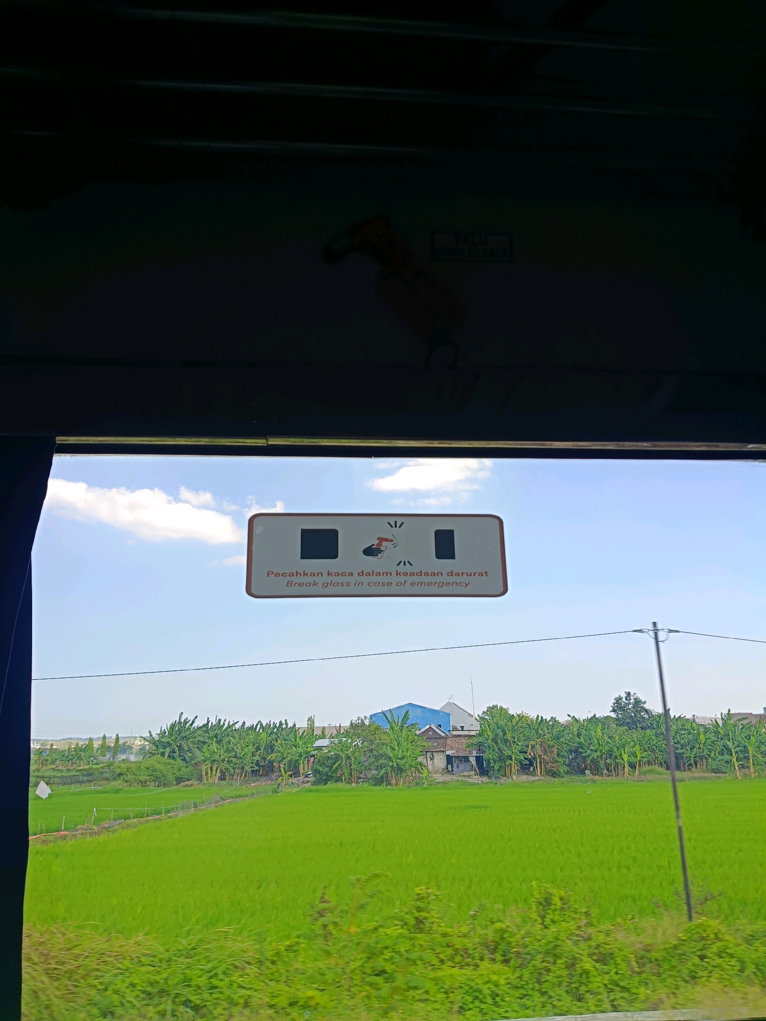 A view from inside a vehicle, looking out a window at a bright, sunny day. A white sign with orange and red accents is mounted on the window. The sign has text that reads "Pecahkan kaca dalam keadaan darurat" and "Break glass in case of emergency" in Indonesian and English, respectively. There are icons on the sign depicting a hammer hitting glass and a hand.
Outside the window, a lush green rice field stretches out to the horizon. Banana trees and a few houses are visible in the distance, interspersed with the greenery. The sky is blue with a few scattered white clouds. A telephone pole with wires stretches across the frame in the mid-ground. The image appears to be taken from a moving vehicle, as evidenced by the slight blur of the background. The overall impression is one of a rural landscape under a clear sky. The city of Sogo, Indonesia is implied as the location.