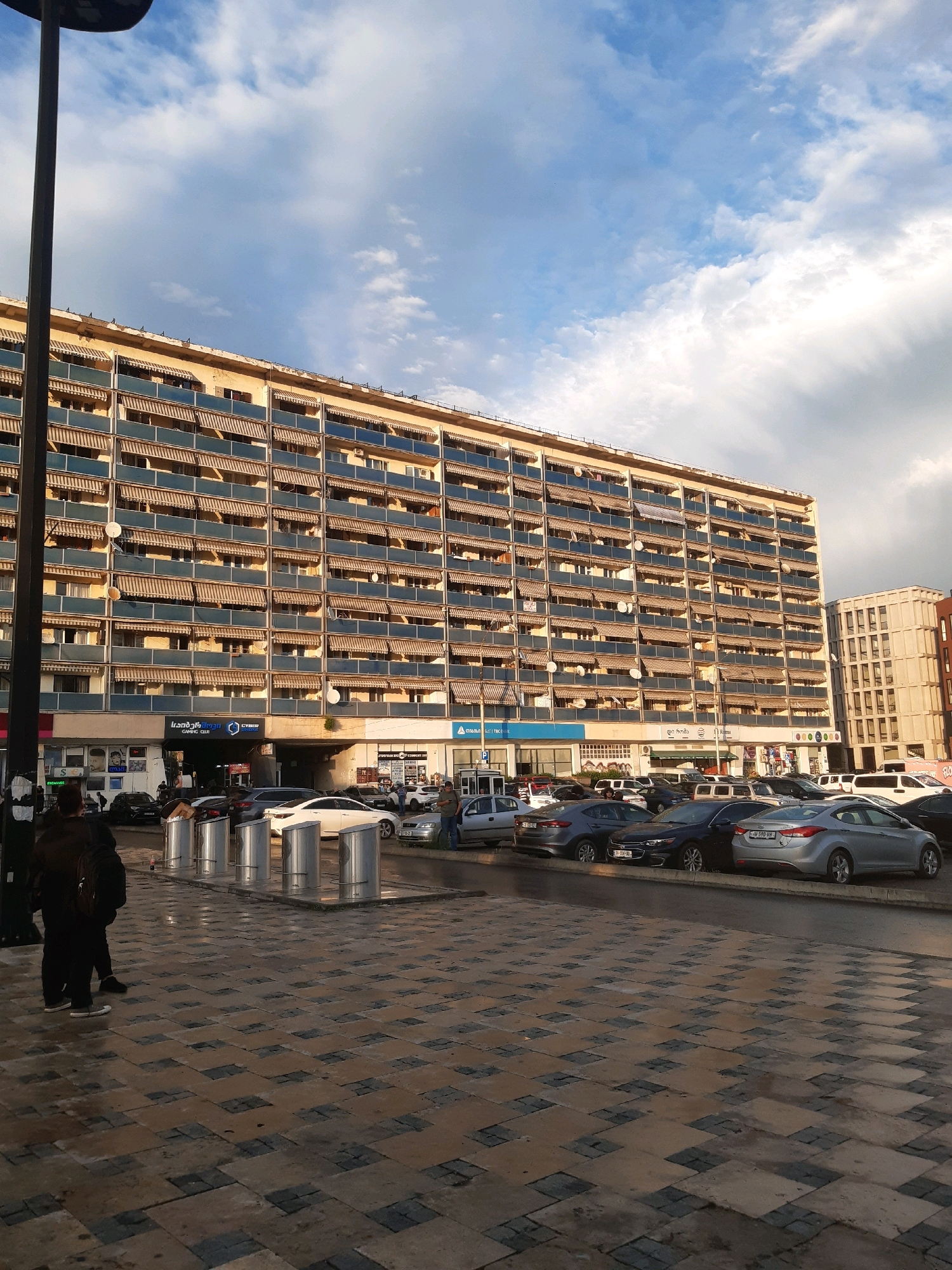 The image depicts a large, multi-story building with numerous balconies, each equipped with a retractable awning. The building is situated in a city environment, with a street in the foreground lined with parked cars. Two individuals are visible in the lower left corner of the image. The sky above is partly cloudy, suggesting a transition in weather. The architectural style and the presence of specific businesses on the ground floor, such as a cyber gaming club and a store with Georgian lettering, indicate the location is Tbilisi, Georgia. The scene is captured in daylight, with shadows cast on the street, indicating the sun's position. The ground is paved with interlocking tiles, some of which appear wet, possibly from recent rain.