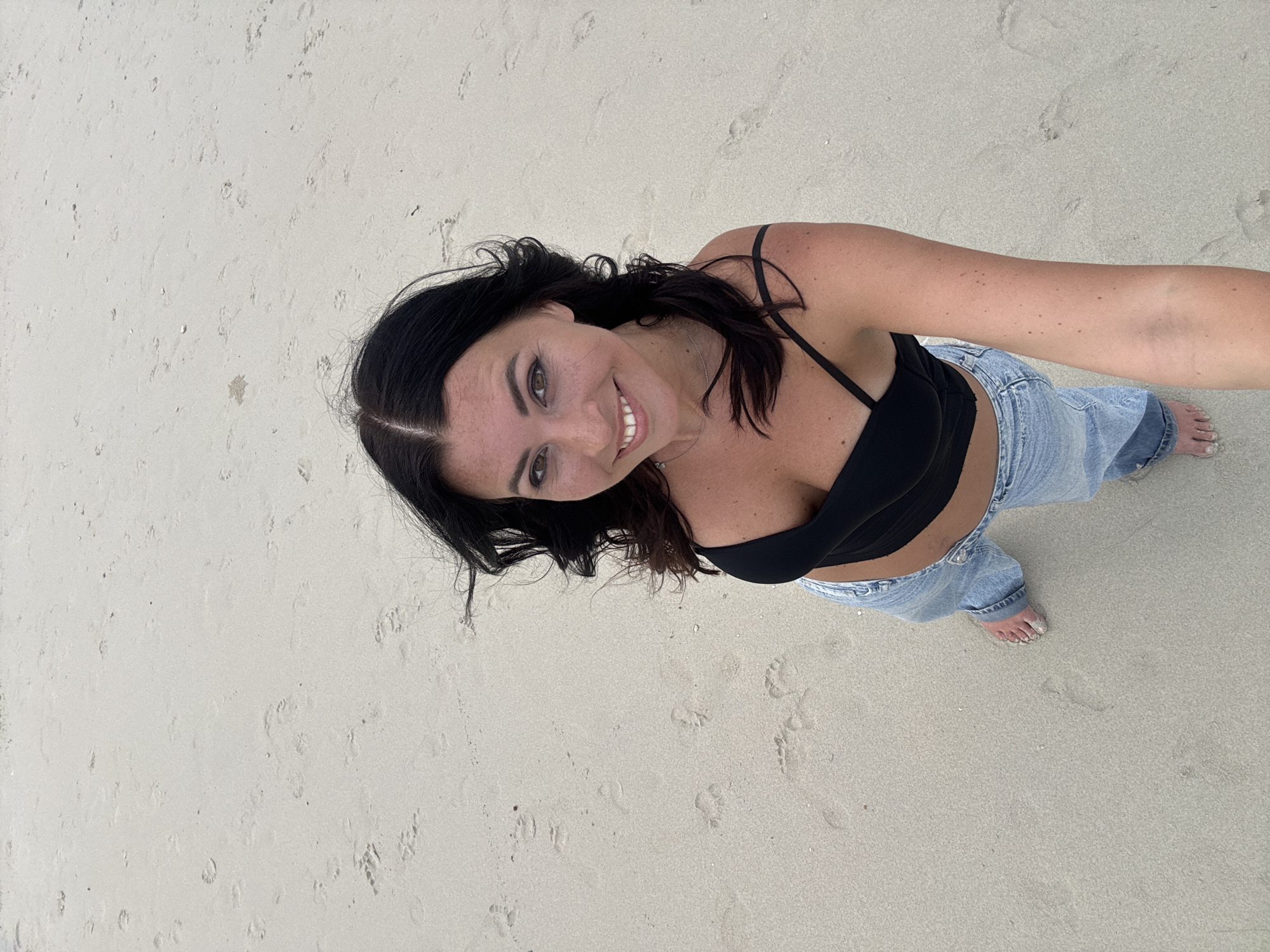 A woman is standing on a sandy beach, possibly in Ban Haad Rin, Thailand. She is wearing a black top and light-wash jeans. Her feet are bare, and there is sand on her toes. The background is a sandy beach. She is smiling and looking up. The photograph is taken from a high angle.