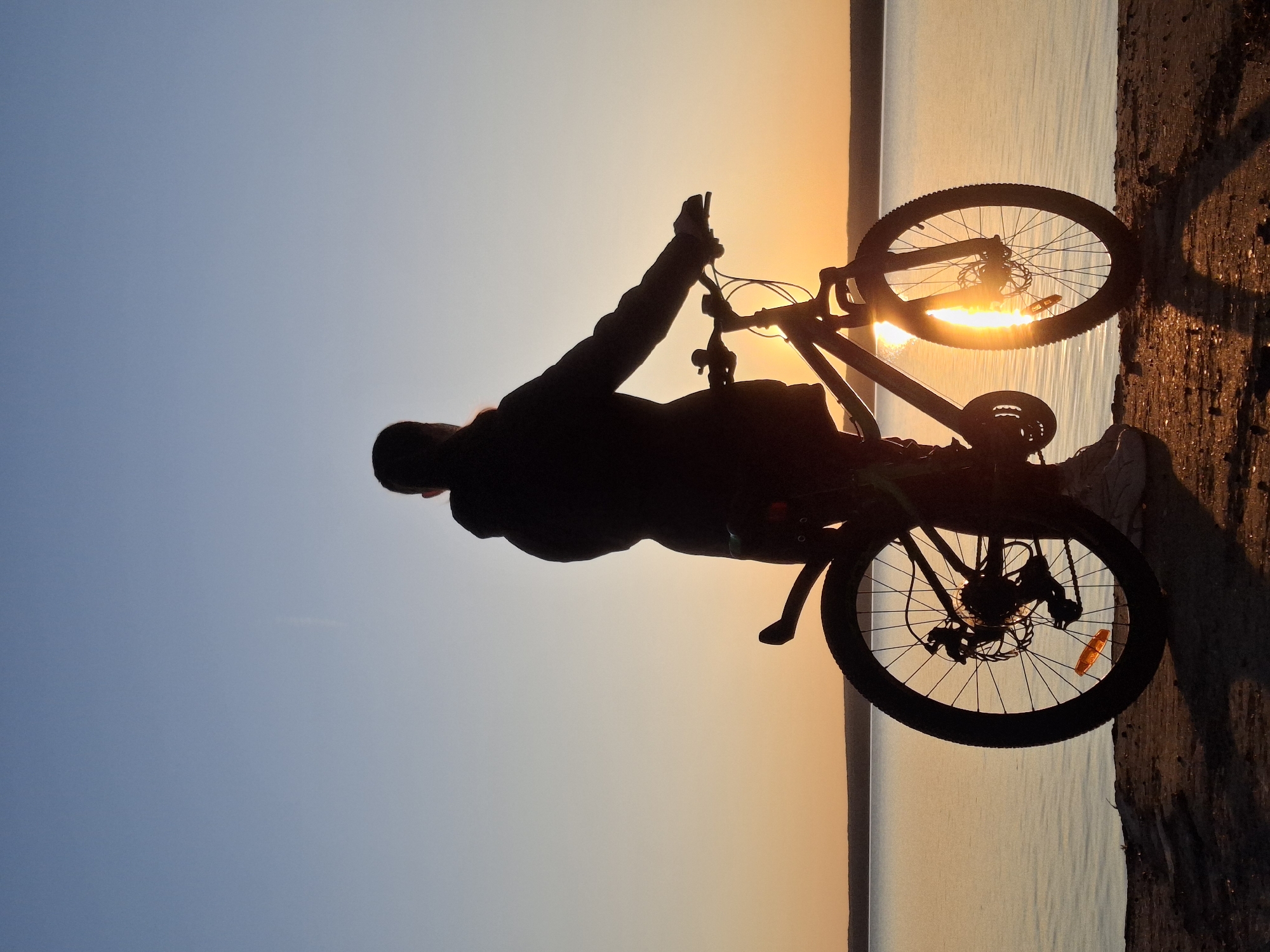 A silhouetted figure stands beside a bicycle, seemingly at sunset, in a location near Chaikovsky, Russia. The light from the sunset is visible through the spokes of the bicycle wheels. The figure appears to be standing on a bank with a body of water visible in the background. The sky has a gradient from a light blue to an orange hue.