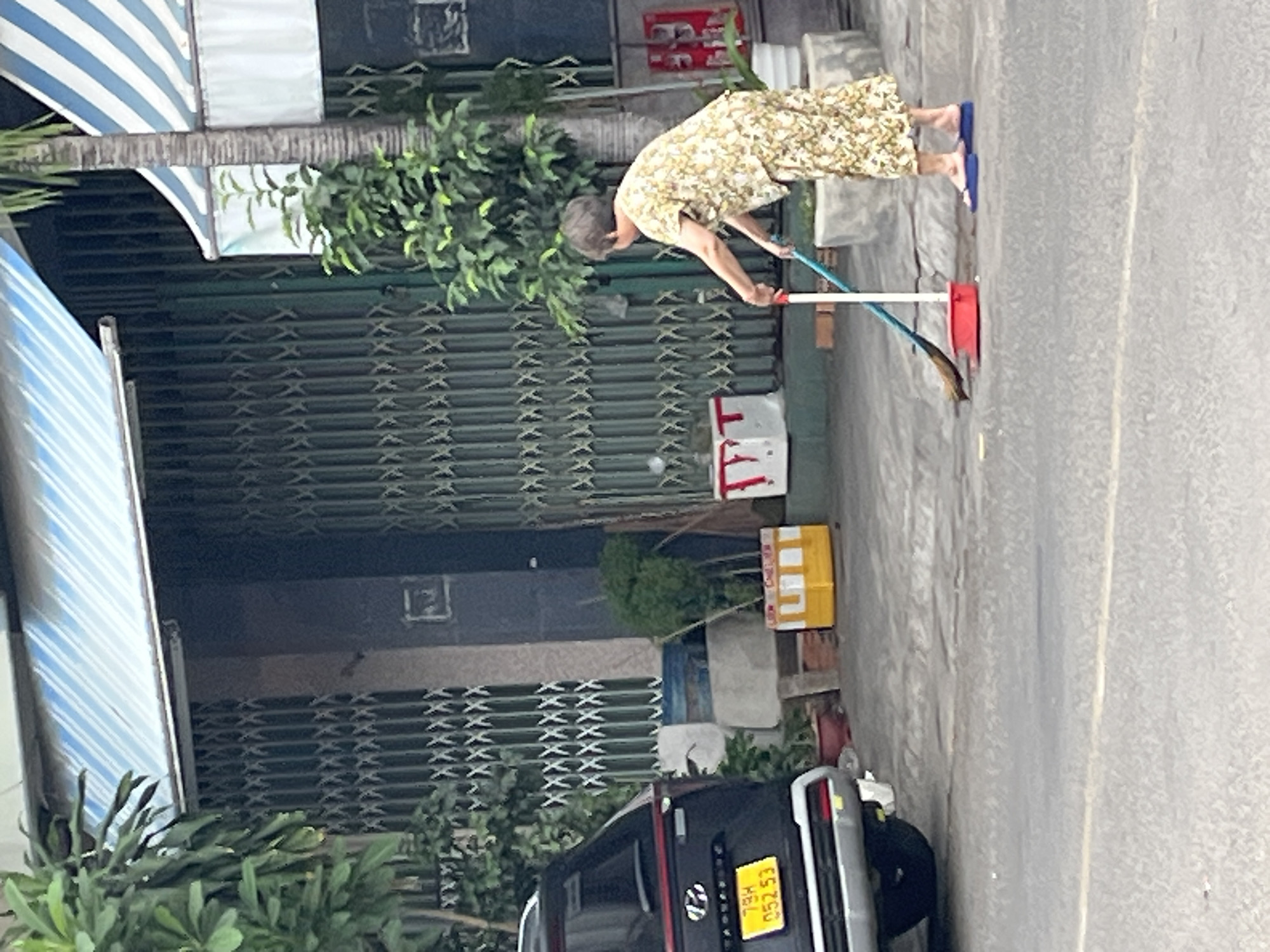 A woman in a floral-patterned outfit and blue flip-flops sweeps the sidewalk in front of a building in Nha Trang, Vietnam. She is using a broom and dustpan. Behind her, a black SUV is parked on the street, and a tall, narrow tree grows next to the building. The building has a corrugated metal awning and security gates. The sidewalk is made of concrete.