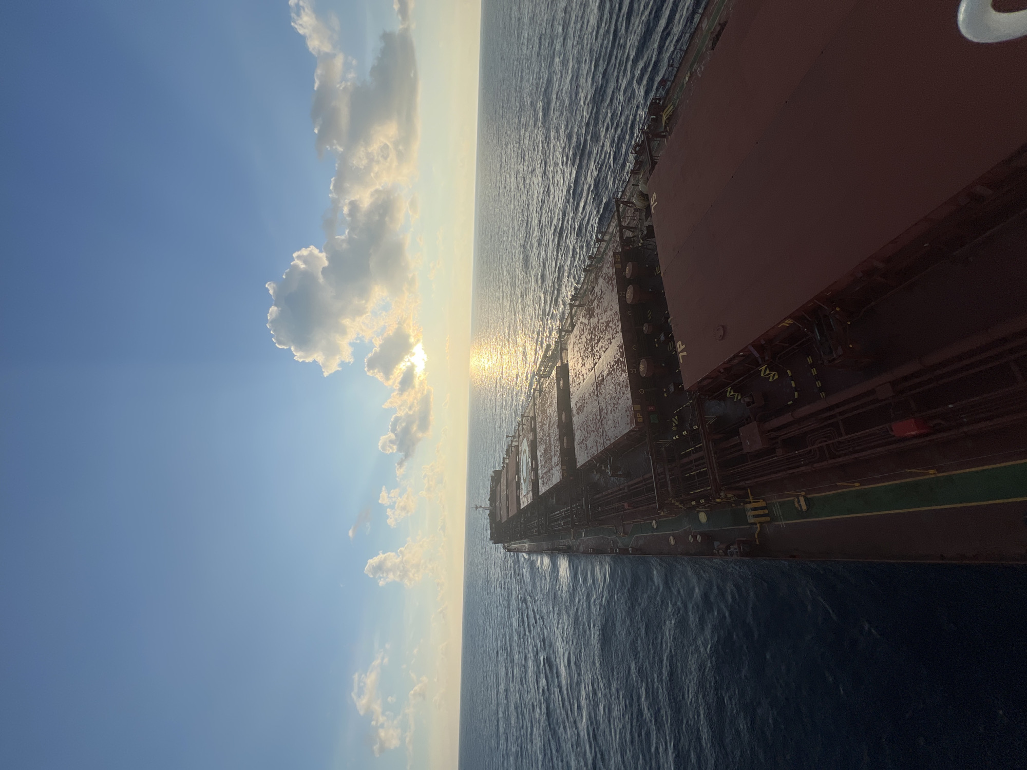 An elevated, high-angle shot captures a large cargo ship sailing on a calm ocean during sunset. The sun, partially obscured by clouds, casts a golden reflection across the water's surface. The ship's deck, a dull red, stretches out towards the viewer, with several large cargo hatches visible. The sky above is a gradient of blue, transitioning to a lighter hue near the horizon where the sun is setting. Scattered white clouds are illuminated by the setting sun, creating a dramatic and serene atmosphere. The ship appears to be moving away from the viewer, indicated by the wake trailing behind it. The water is a deep blue, with gentle ripples suggesting minimal wave activity. The overall scene is one of peaceful travel at sea during a beautiful sunset.