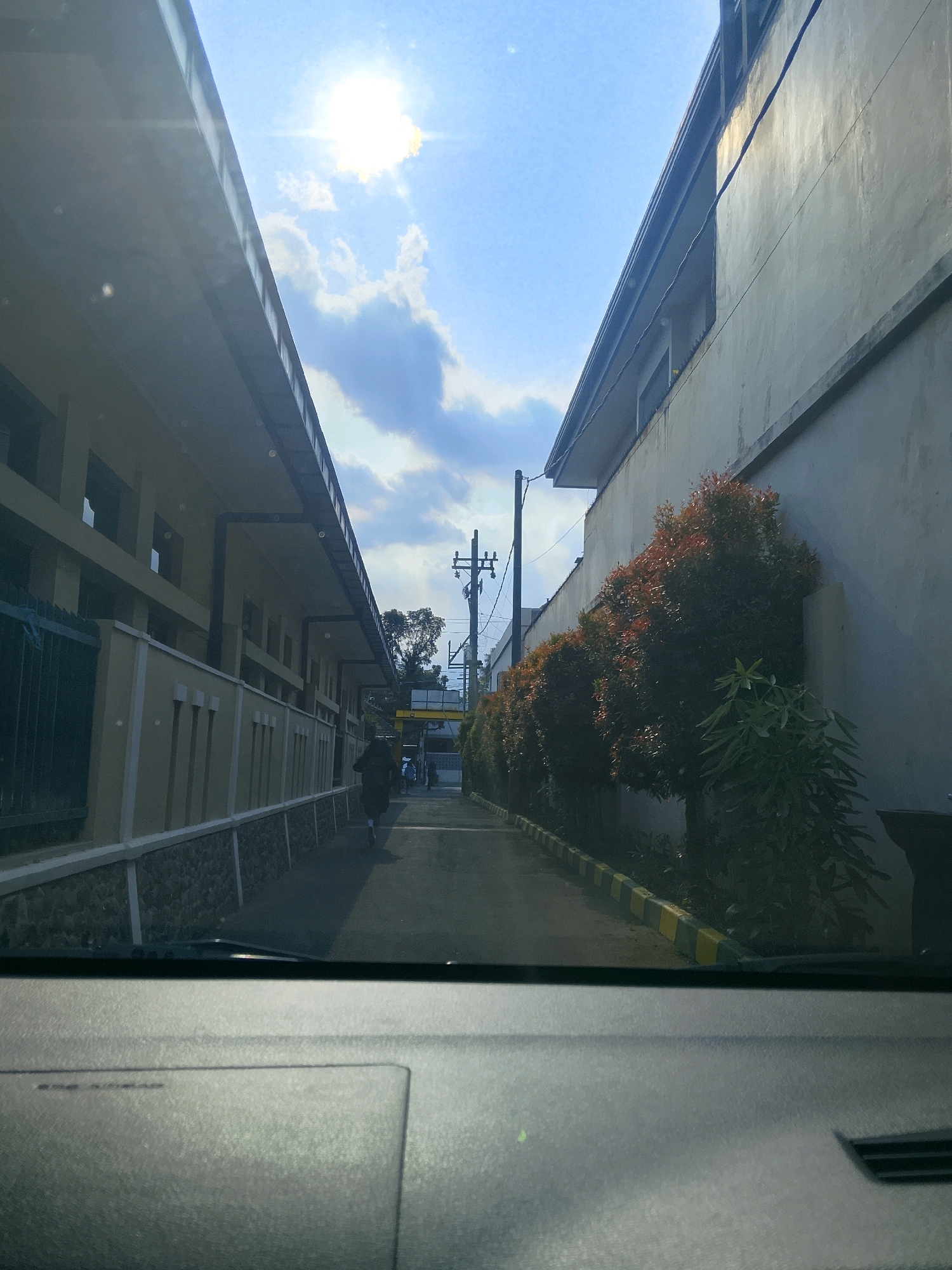 The image shows a view from inside a vehicle, looking forward through the windshield. The perspective suggests the vehicle is moving down a narrow street or alleyway lined with buildings on both sides. The building on the left is lighter in color and appears to be a low-rise structure with a covered walkway. The building on the right is a taller, more solid structure with a concrete-like exterior.
Along the right side of the alley, a row of trees with red-tinged foliage is planted. The ground is paved, with yellow and green striped curbs visible along the edge of the sidewalk or pathway. In the distance, a person is walking away from the camera. Further down the alley, there's a utility pole with electrical wires and what appears to be a yellow overhead structure. The sky is bright blue with scattered clouds, and the sun is visible, casting a glare.
The image is captured during daylight. The location is described as being in the city of Malang, Indonesia. The scene depicts a typical urban environment, possibly a residential or commercial area with a focus on pedestrian and vehicular traffic. The overall impression is one of a bright, sunny day in a somewhat institutional or well-maintained environment.