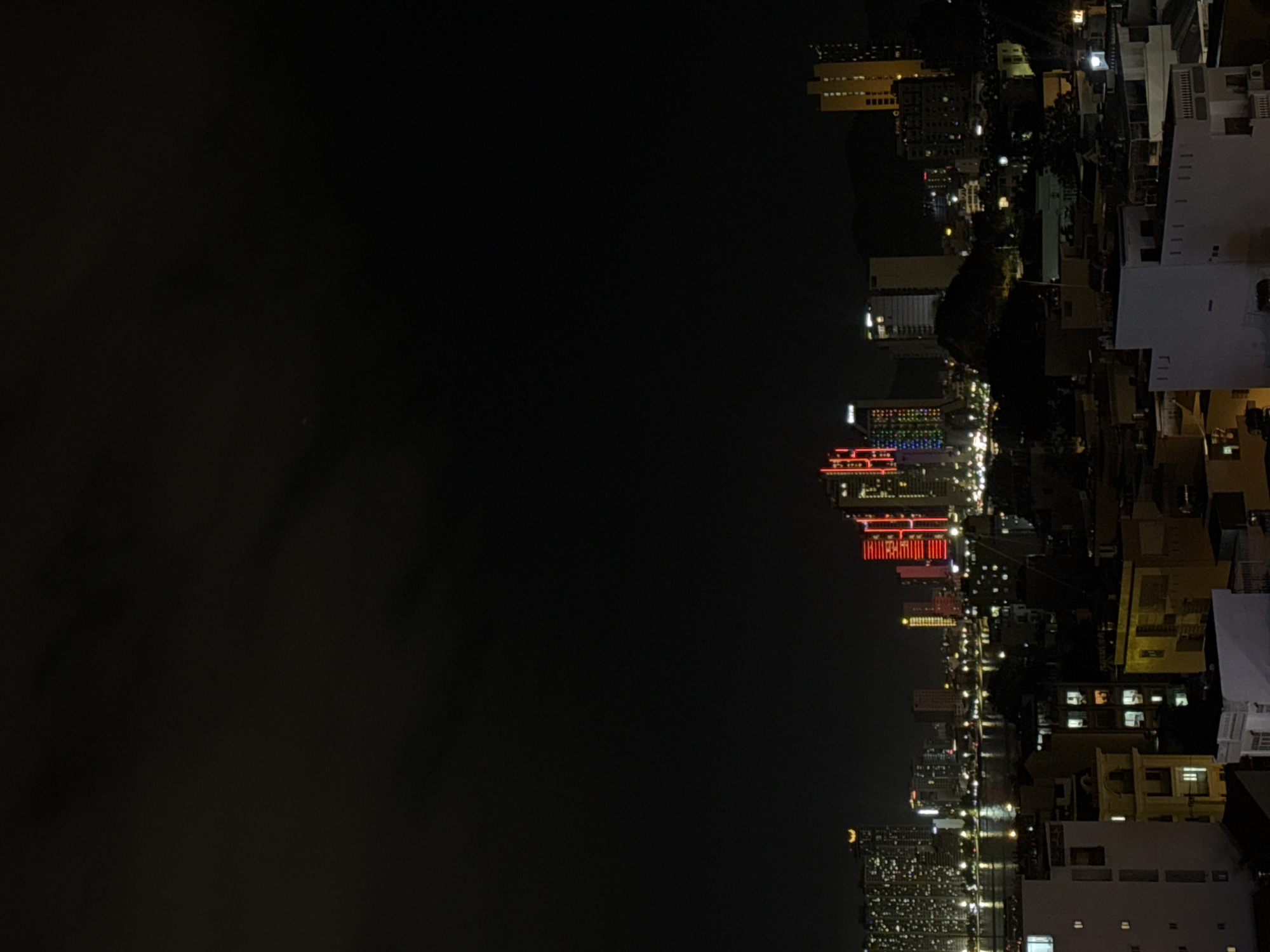 The image captures a nighttime cityscape in Bac Nha Trang Ward, Vietnam, characterized by a dense urban environment with a prominent waterfront. The foreground consists of several low-rise buildings, some illuminated with interior lights, suggesting residential or commercial occupancy. Beyond these structures, a body of water, likely the sea or a bay, is visible, reflecting the lights of the city and a distant bridge.
The middle ground is dominated by a cluster of taller buildings, including several skyscrapers. Some of these taller structures are illuminated with vibrant, colorful LED lights, predominantly in red and multi-colored patterns, creating a striking visual contrast against the dark sky. One notable building features a distinct golden crown or architectural element at its apex.
The background features a dark, cloud-filled sky, characteristic of nighttime conditions. The silhouette of mountains or hills is faintly visible in the far distance. The overall impression is of a bustling, modern coastal city at night, with a strong emphasis on illuminated architecture. No people or specific actions are discernible due to the distance and lighting conditions. The scene is presented from an elevated viewpoint, looking out over the city.