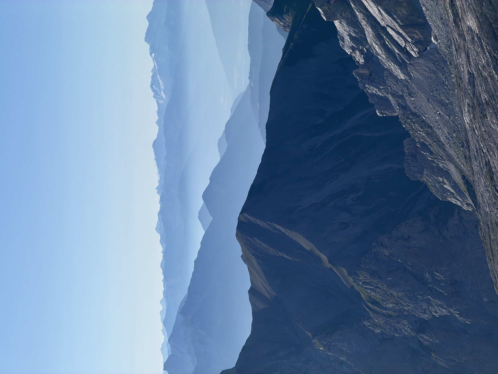 The image depicts a panoramic view of the Bavarian Alps near Garmisch-Partenkirchen, Germany. The scene is characterized by layers of mountain ranges receding into the distance, creating a sense of depth and scale. The foreground is dominated by rugged, dark gray rock formations, indicative of a high-altitude, rocky terrain. Further back, progressively lighter blue hues define successive mountain ridges, softened by atmospheric haze. The distant peaks are barely visible, appearing as faint silhouettes against a clear, pale blue sky. The sunlight suggests it is daytime, likely morning or late afternoon, casting subtle shadows and highlighting the contours of the mountains. There are no people, structures, or visible signs of human activity in the image, emphasizing the natural, unpopulated landscape. The environment is pristine and wild, showcasing the grandeur of alpine scenery.