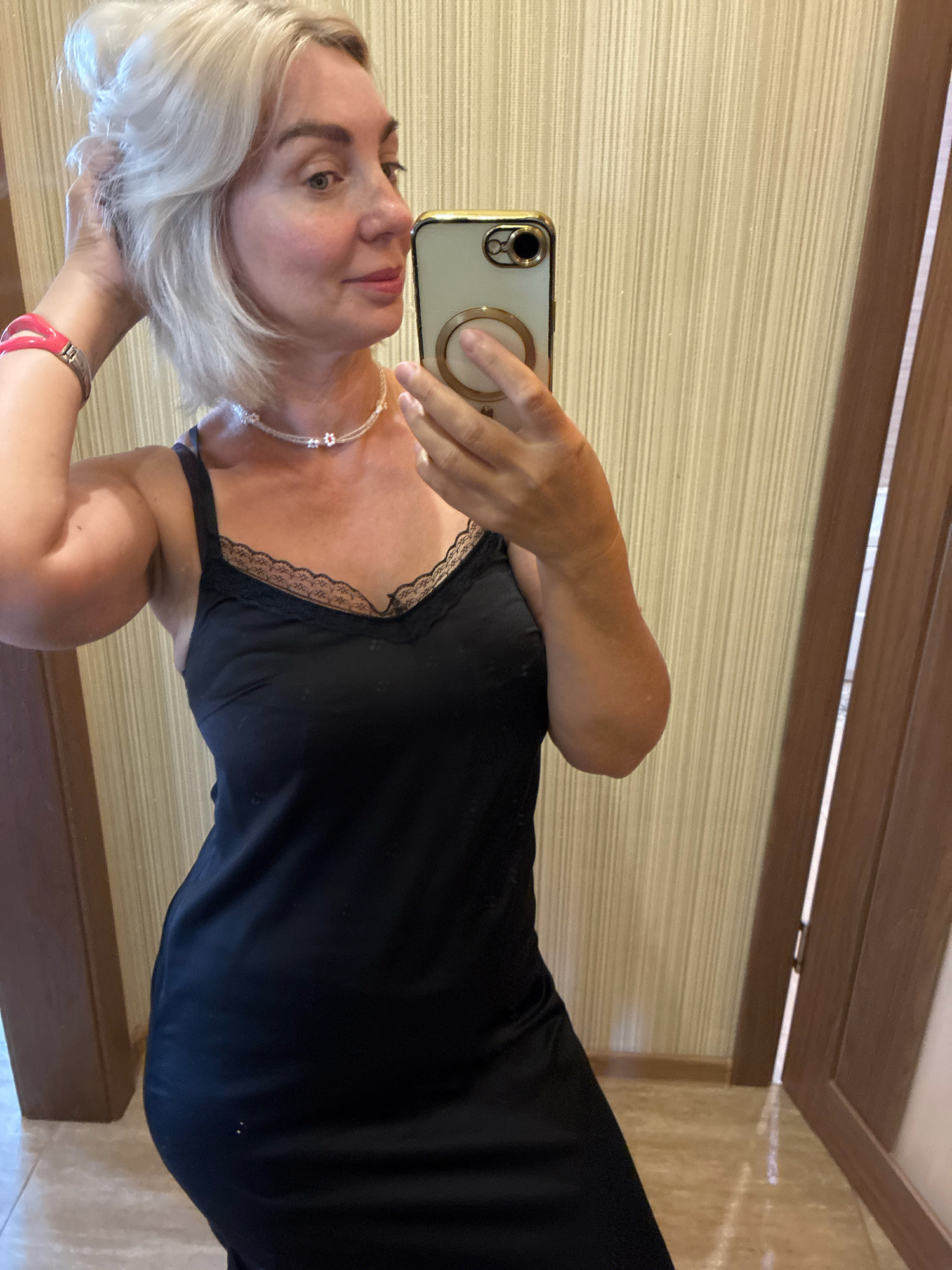 An adult Caucasian woman with blonde hair and fair skin is taking a mirror selfie. She is wearing a black slip dress with lace trim and a beaded necklace. A pink bracelet is visible on her left wrist. She is holding a smartphone in her right hand, which is angled to capture her reflection. The background appears to be an indoor setting with textured wallpaper and a doorway. The image does not contain information that places it in Sochi, Russia.