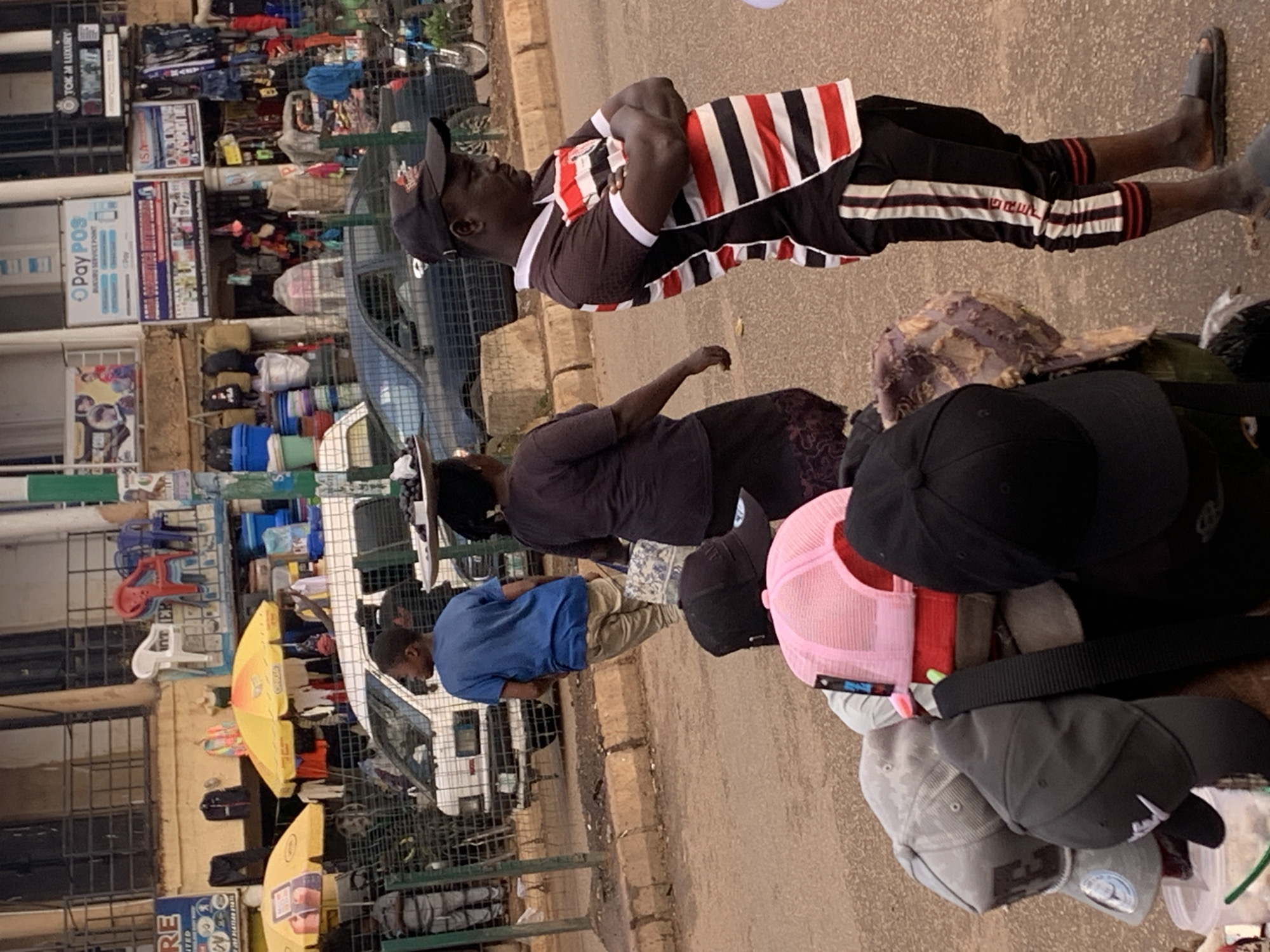 This image depicts a bustling market scene in Bukuru, Nigeria. The primary subjects are several people, including a man standing with his arms crossed, wearing a black, red, and white striped shirt and black shorts. Another individual, a woman, is partially visible behind him, wearing a dark shirt and patterned skirt. In the foreground, there are several caps, including a pink mesh trucker hat, black baseball caps, and a grey cap. The scene is set on a street next to market stalls, with metal grates, shelves filled with goods like clothing and household items, and a white vehicle visible. Yellow umbrellas are set up, suggesting a vendor's area. The lighting indicates it's daytime, and the overall atmosphere suggests a lively commercial environment. Visible text includes "PayPOS" on a sign for a service point and potentially other vendor signage that is not clearly legible. The image is oriented vertically.