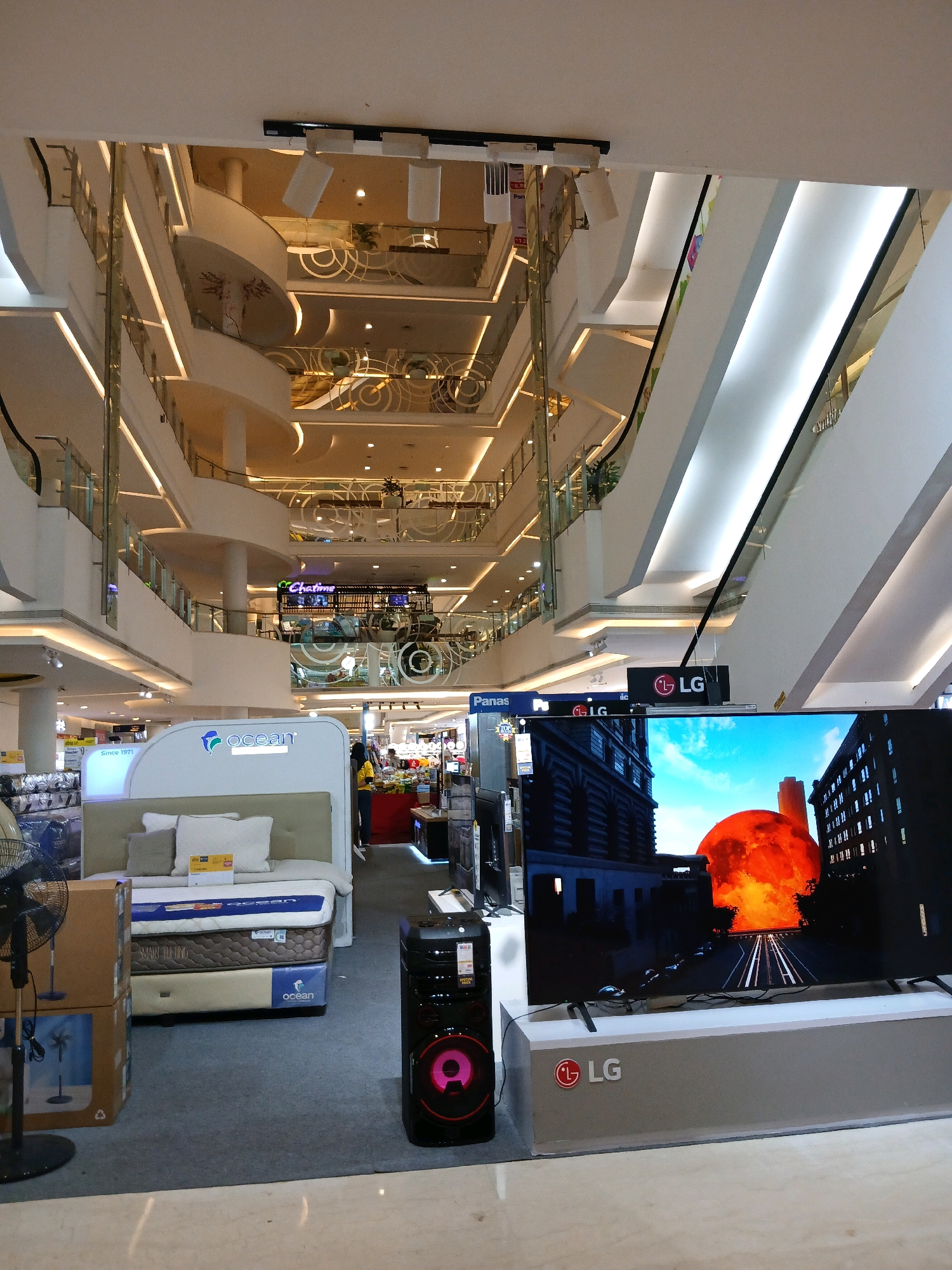 The image depicts the interior of a modern shopping mall in North Jakarta, Indonesia, likely an electronics store. In the foreground, a large LG television displays a dramatic image of a large orange celestial body or explosion over a city skyline. To the left of the TV is a black speaker with a pink illuminated woofer. Further left, a display bed with pillows is set up, branded with "ocean". A black standing fan is positioned in front of stacked cardboard boxes. The background reveals multiple levels of the mall with escalators, glass railings, and ambient lighting, suggesting a spacious and multi-tiered structure. Several retail signage, including "Panas" and "LG," are visible on the upper levels. A person in a yellow shirt is briefly visible in the mid-ground.