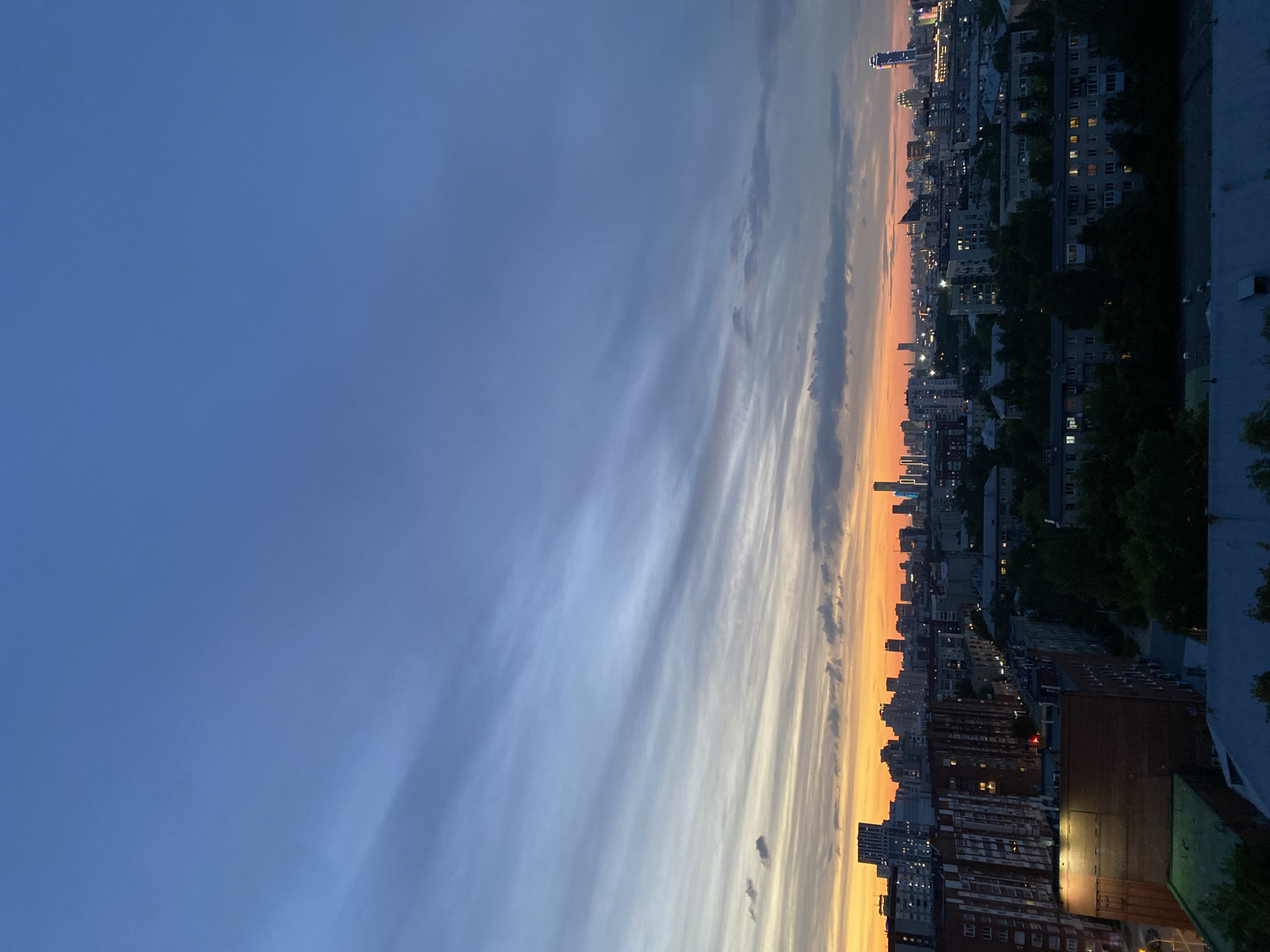 The original image presents a panoramic view of the Yekaterinburg, Russia cityscape, captured during either dusk or dawn and oriented in portrait mode. The perspective is from a high vantage point, likely a building or rooftop.
The sky dominates the left side of the frame, exhibiting a striking color gradient. It begins with a deep, clear blue at the top-left, gradually transitioning through lighter blues and grays, and culminates in vivid oranges and yellows along the right edge, indicating the position of the sun near the horizon. Long, streaky cloud formations stretch horizontally across the middle section of the sky, illuminated by the sun's light to display a mix of white, gray, and orange hues.
The right side of the image showcases the dense urban landscape. Numerous multi-story buildings, varying in height and style, form a silhouette against the brightly lit horizon. Many of these structures display illuminated windows, suggesting that interior lights are on, reinforcing the impression of evening. Scattered trees are visible among the buildings, adding dark green accents to the scene.
A notable feature in the upper-right portion of the skyline is a tall, slender building with a distinct spire-like top, catching the last warm light. In the immediate foreground, at the bottom-right, parts of rooftops are visible, including one with a green-patinated surface. The overall scene evokes a sense of peaceful urban transition, with the city lights beginning to emerge against the beautifully colored sky. No human activity or visible text is discernible in the image.