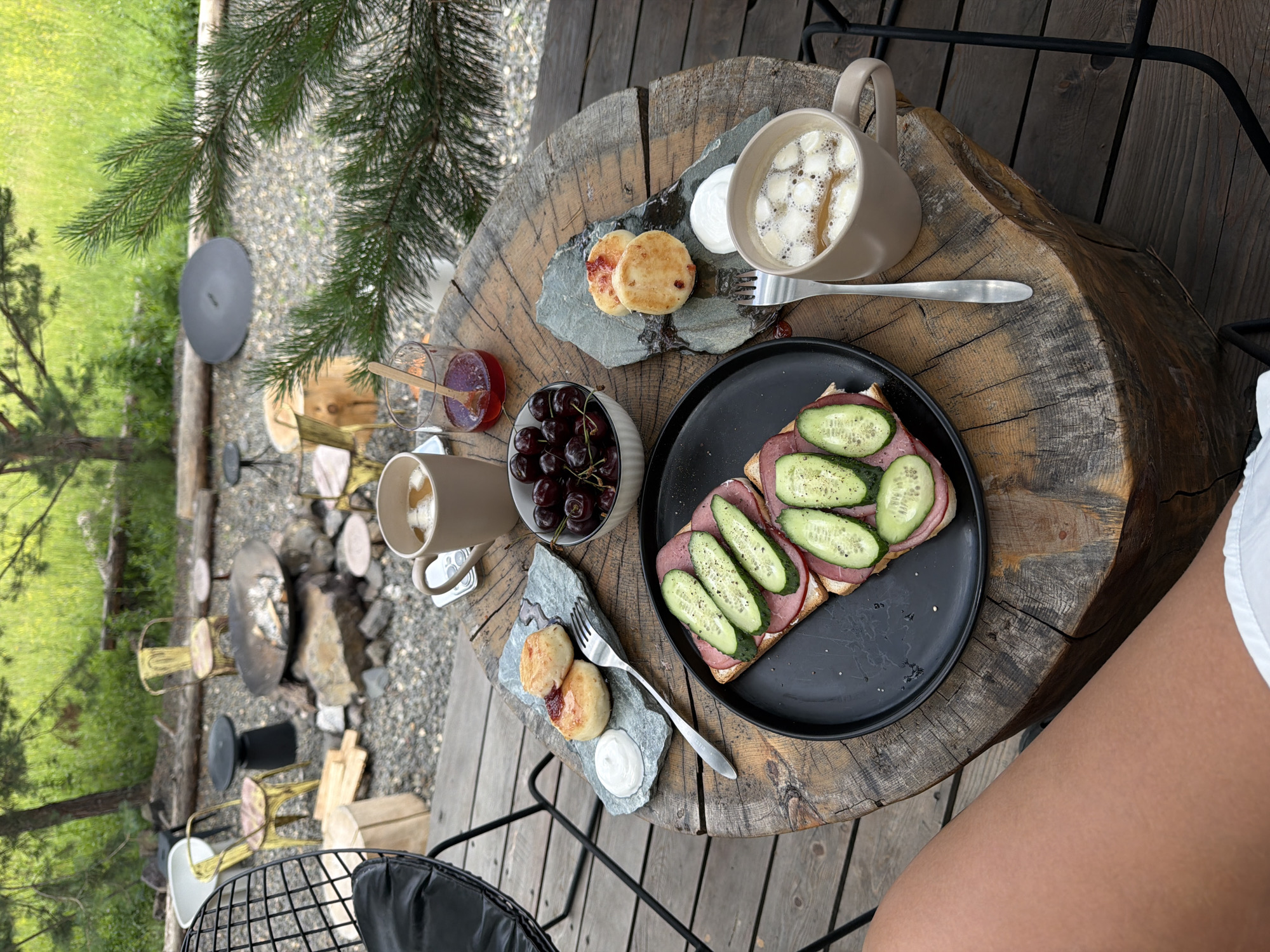 A rustic outdoor setting, possibly a campsite or a lodge in or near the city of Mansky, Russia, features a wooden table laden with breakfast items. The table, appearing to be a large tree stump, is set with a black plate holding a sandwich topped with ham and sliced cucumbers, a small bowl of cherries, and two gray slate plates each with two small pastries and a dollop of cream. A fork rests on one slate plate, and another fork is near a mug of coffee. A glass with a spoon and jam sits beside the cherries. In the background, a fire pit area with rocks and chairs is visible, surrounded by greenery and trees. A person's legs are visible in the foreground, suggesting they are seated at the table.