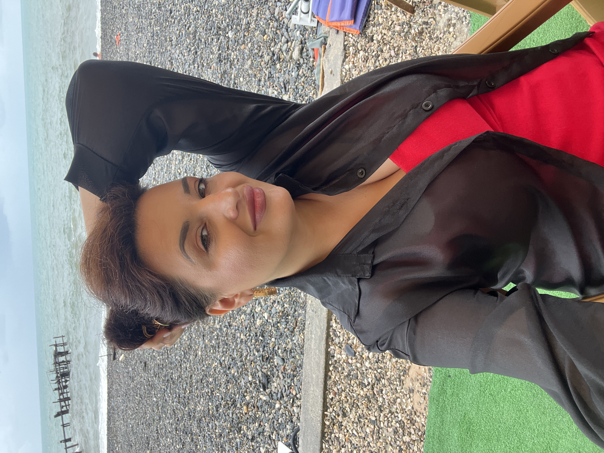 The image shows a woman from the chest up, posing for a selfie at a beach in Sokhumi, Abkhazia. She is wearing a black open shirt over a red dress. Her hair is styled in a bun on top of her head, and she has large gold hoop earrings. The background shows a pebble beach, the Black Sea, and a cloudy sky. There are some wooden pilings in the water, and a few people are visible in the distance on the beach. The woman is sitting on a chair, with a portion of a green artificial turf visible in the lower left corner.