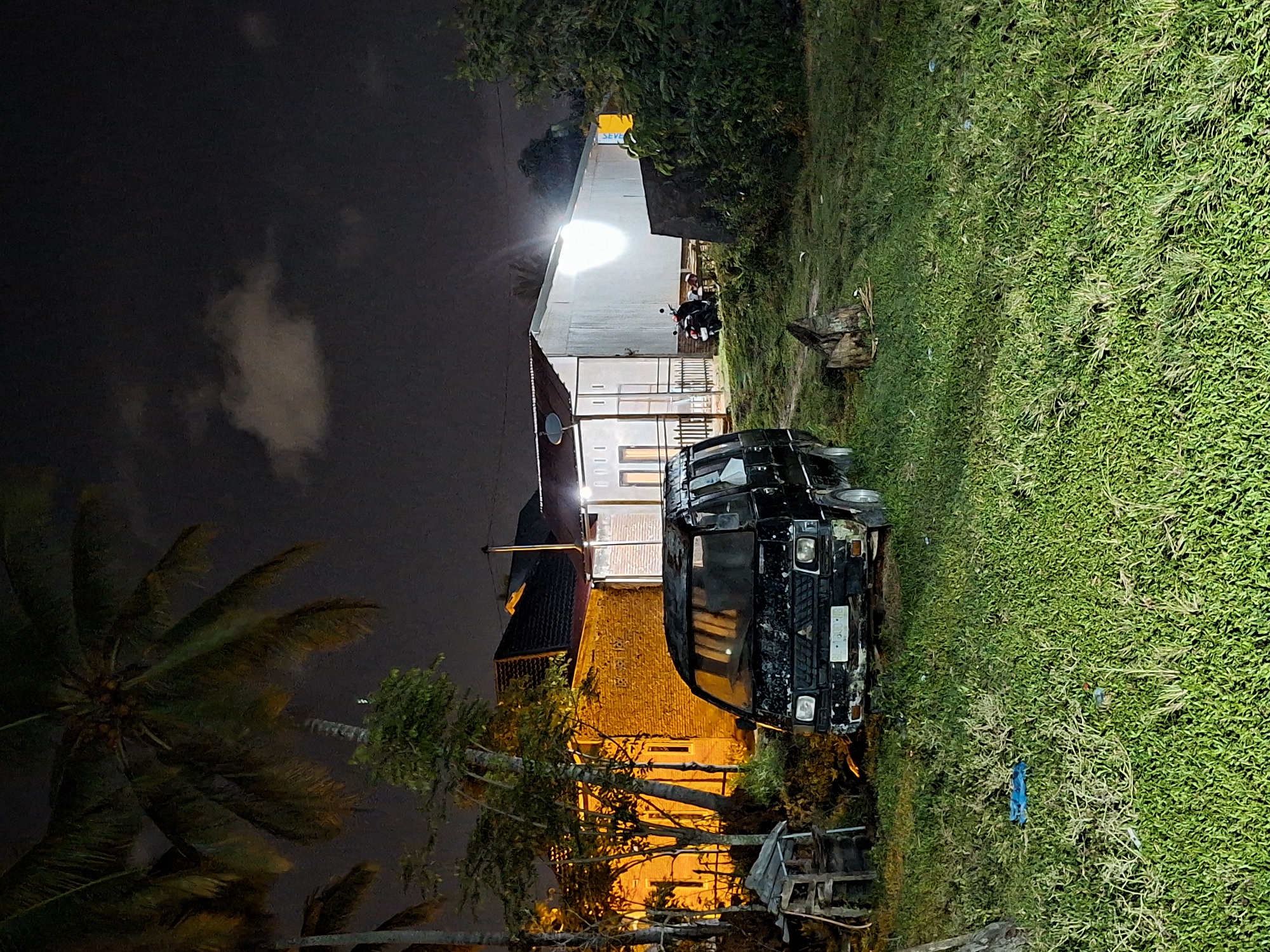 The image depicts an outdoor scene at night in Lhokseumawe, Indonesia. A heavily damaged black van is parked on a grassy area next to a building. The building appears to be a residence, illuminated by warm light from within and a bright security light above. A motorcycle is parked nearby. A palm tree is visible in the foreground, and a cloudy night sky forms the backdrop. Debris and old furniture are scattered near the vehicle.